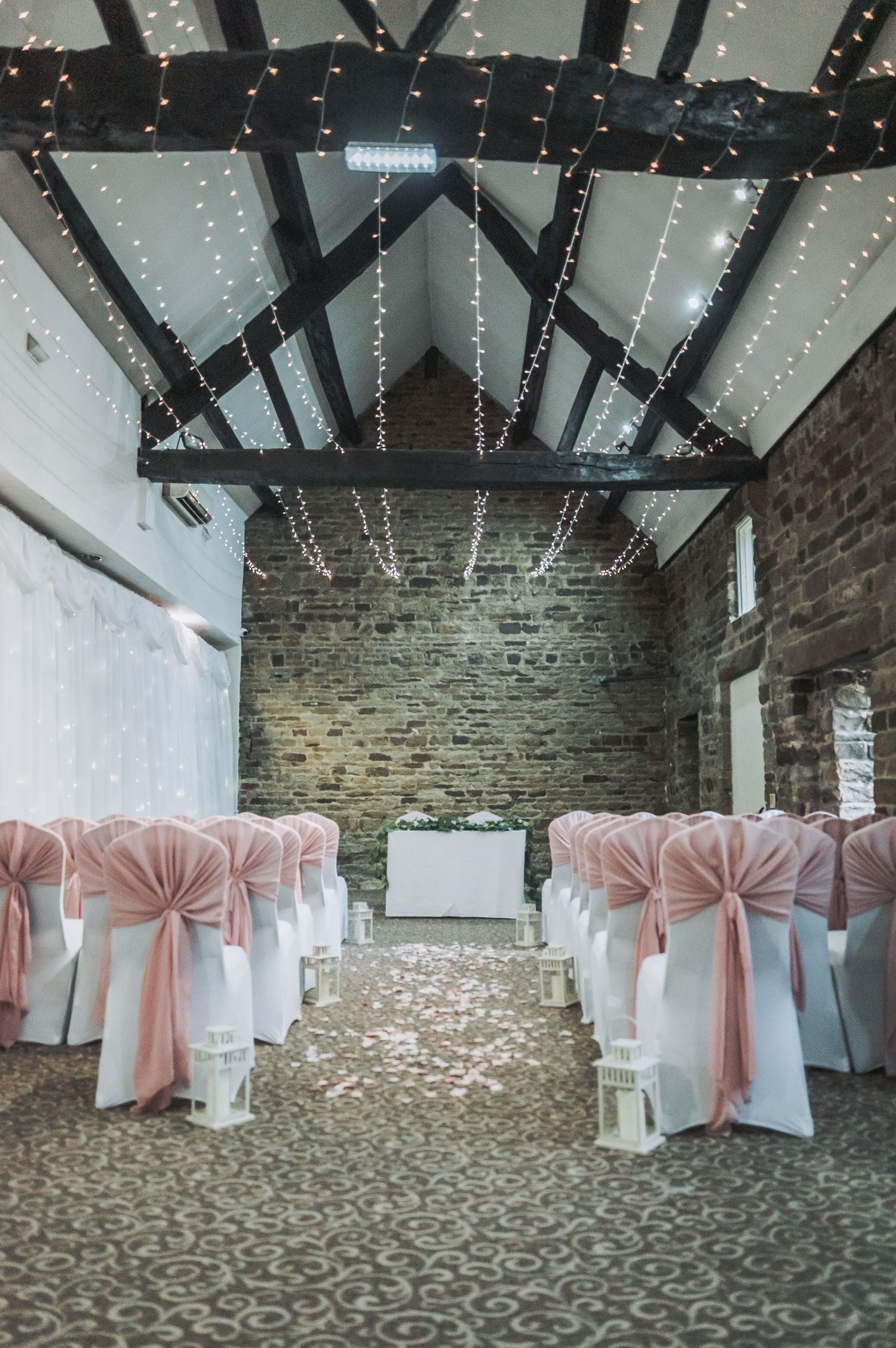 Wedding ceremony venue with white chairs, pink sashes and fairy lights decorating exposed wooden beams.