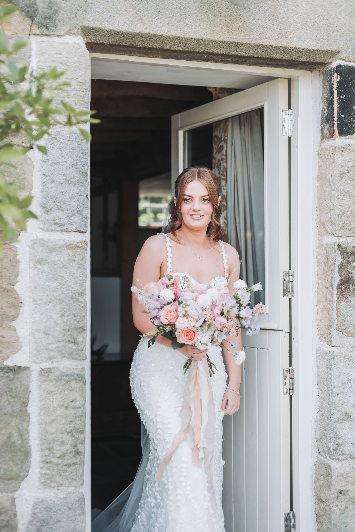 Bride with bouquet stands in doorway of stone building wearing lace wedding gown.
