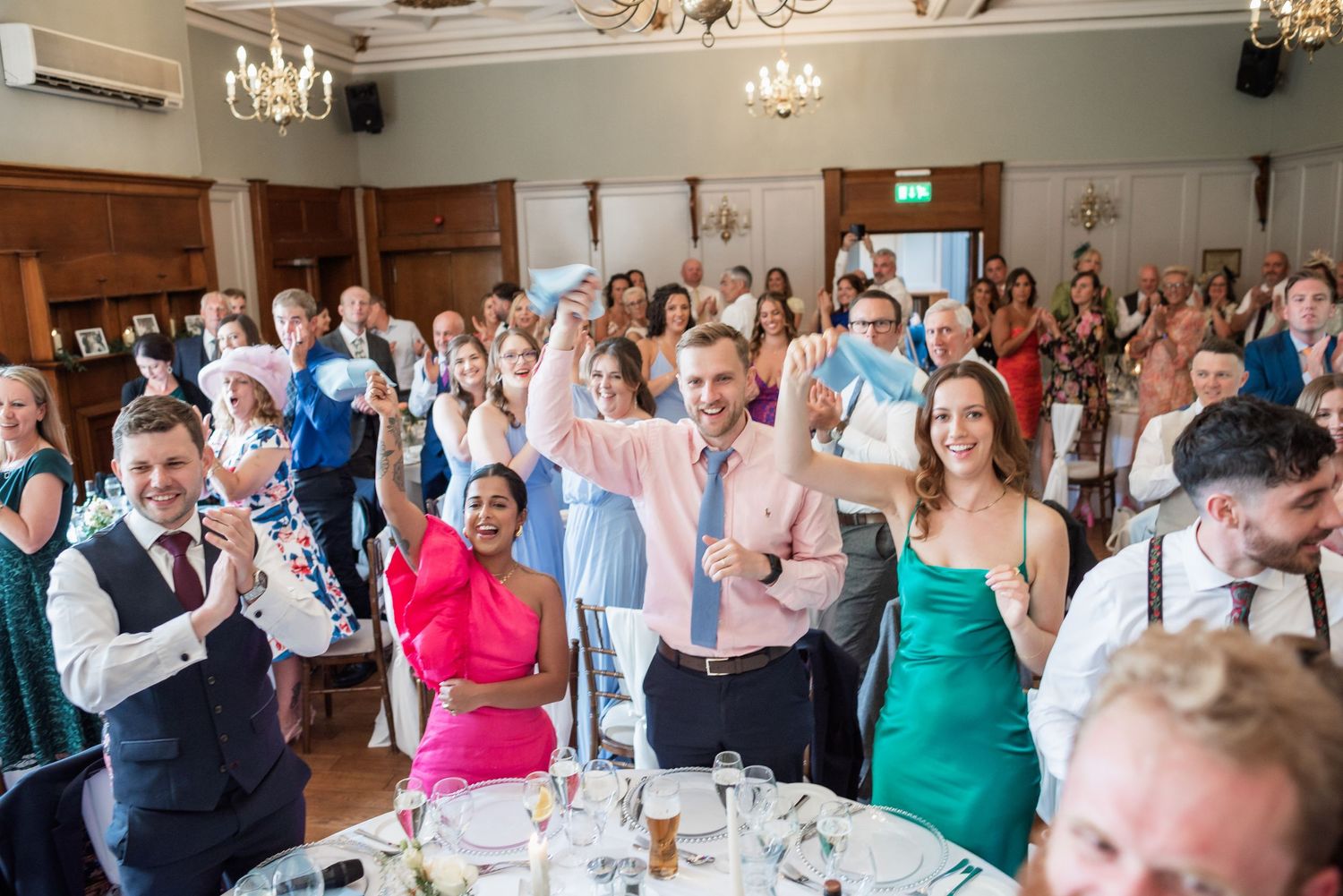 Wedding reception guests cheering and celebrating in an elegant ballroom with chandeliers.