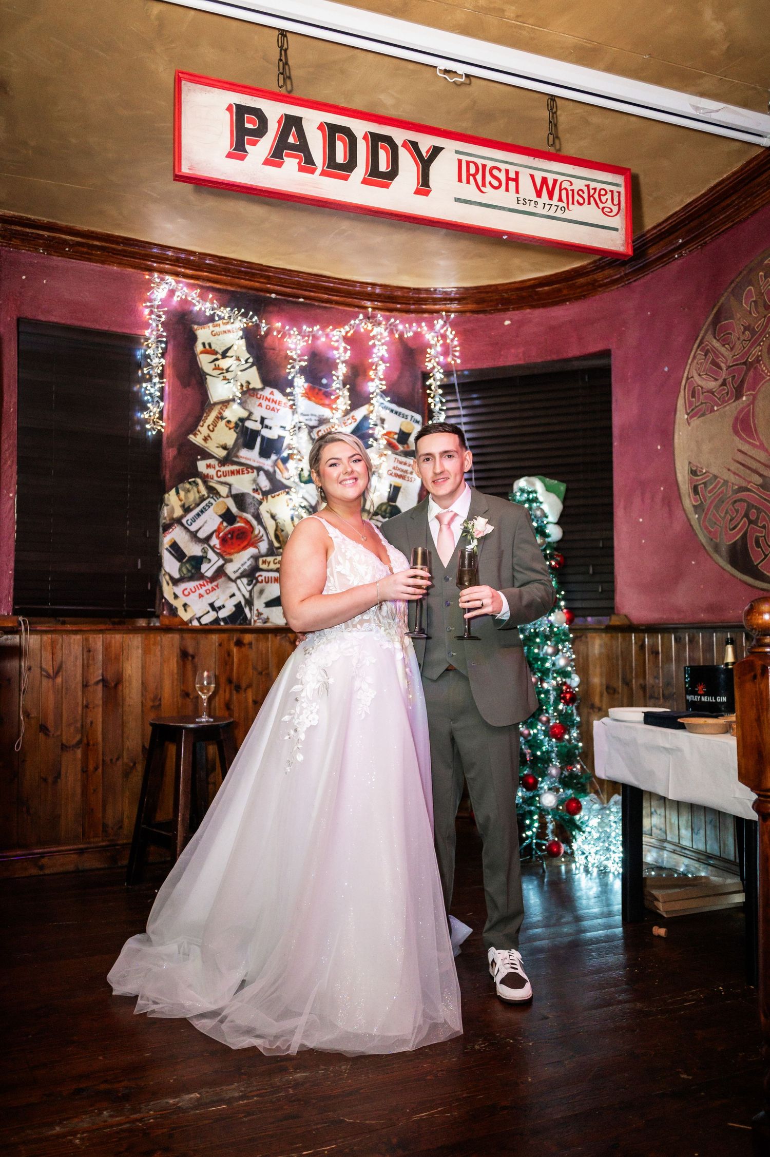 Newlyweds pose with drinks under Paddy Irish Whiskey sign in pub setting.