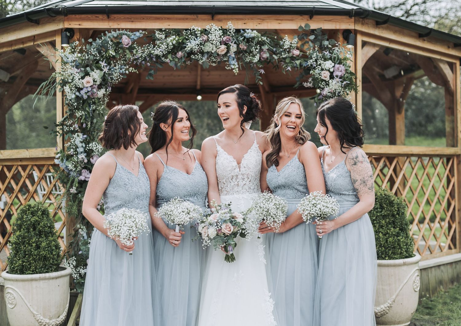 Bridesmaids in pale blue dresses pose with bouquets by a gazebo.