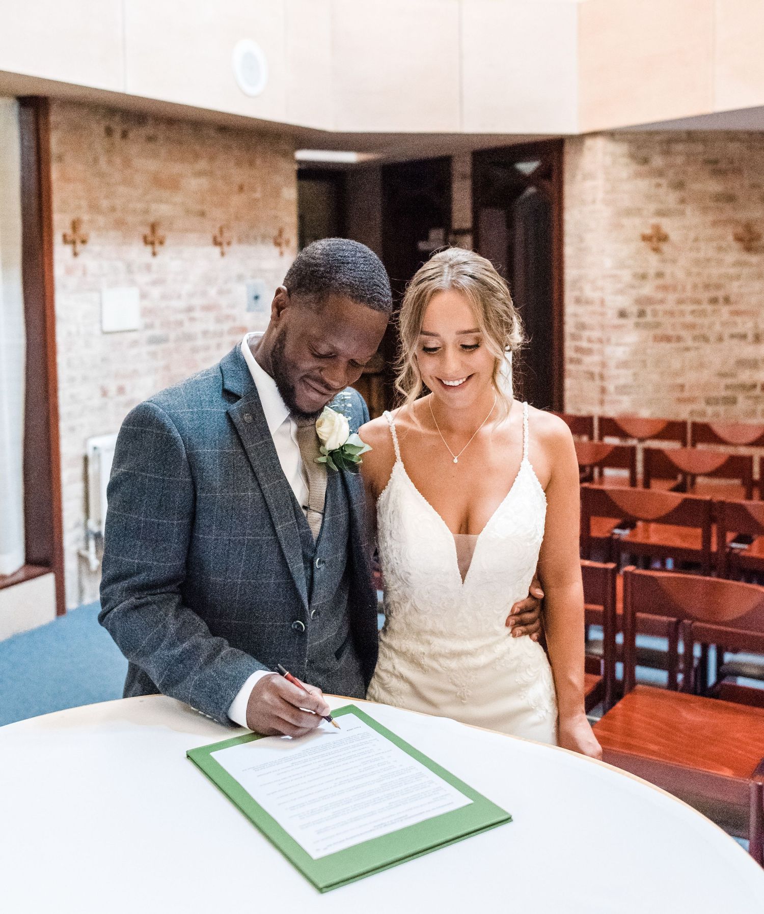 A couple signs their marriage license together at a table in a brick-walled church.