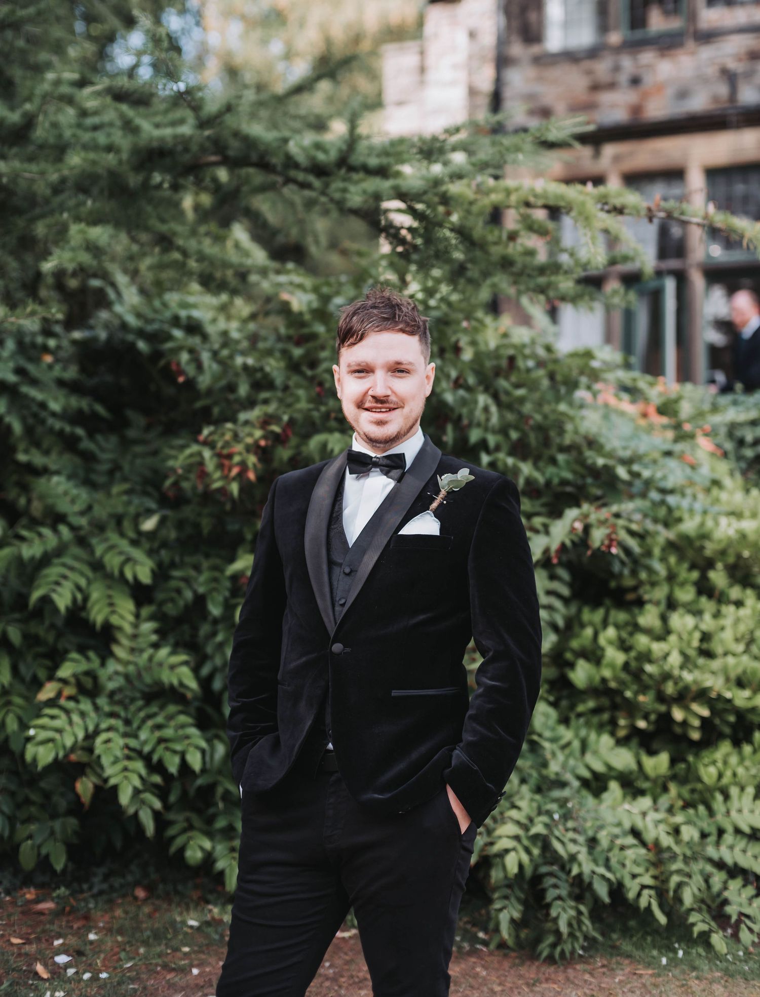 A groom in a black velvet tuxedo and bowtie poses outdoors against greenery at a wedding venue.