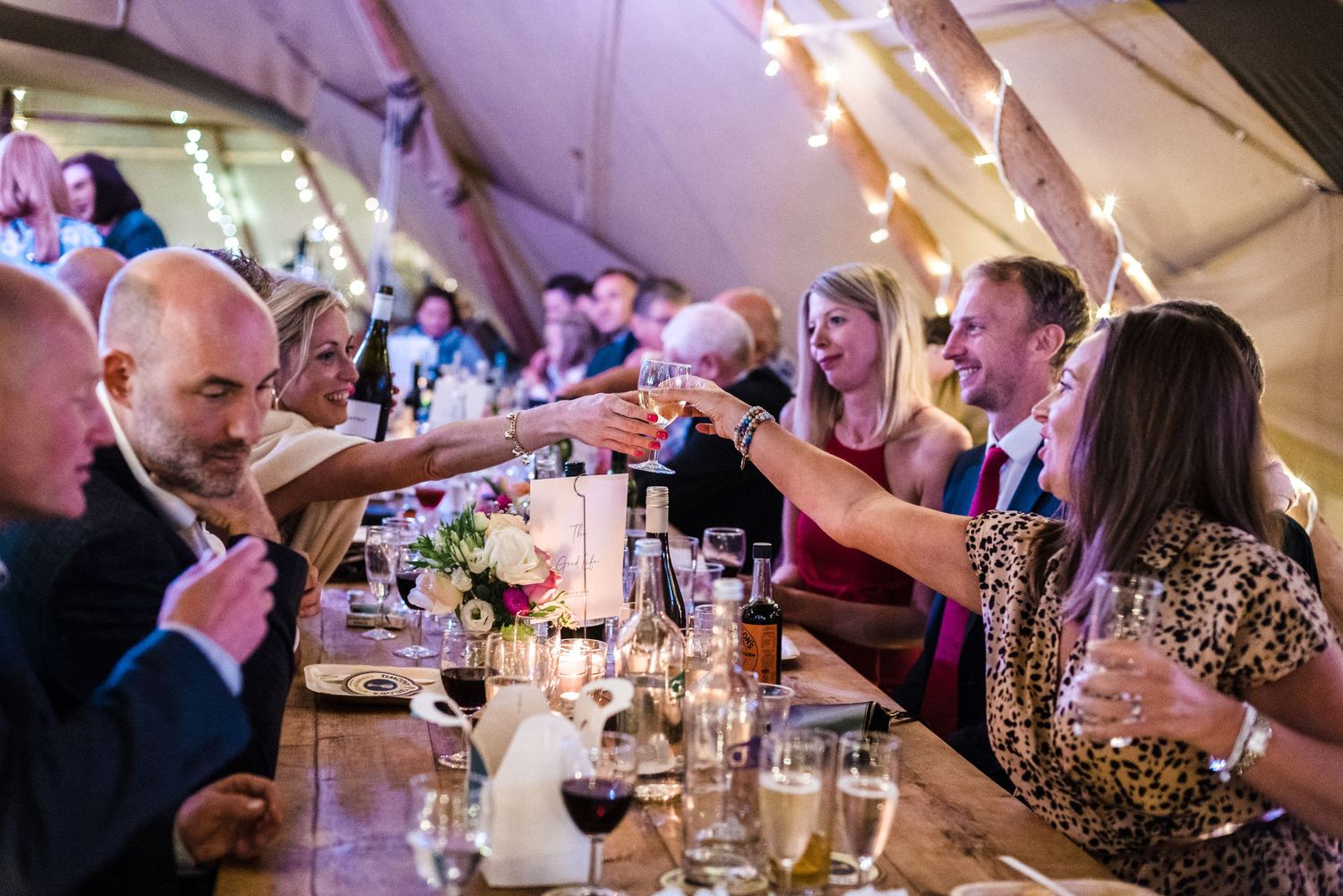 Wedding guests toasting and celebrating at a long dining table under string lights in a tent reception.