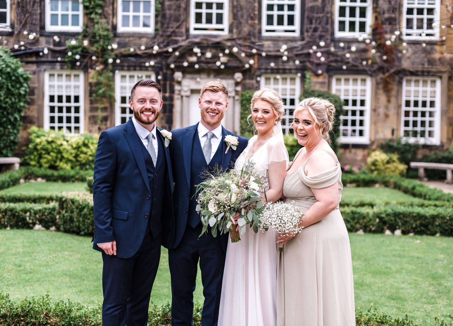 A wedding group poses in formal attire against a historic ivy-covered building backdrop.