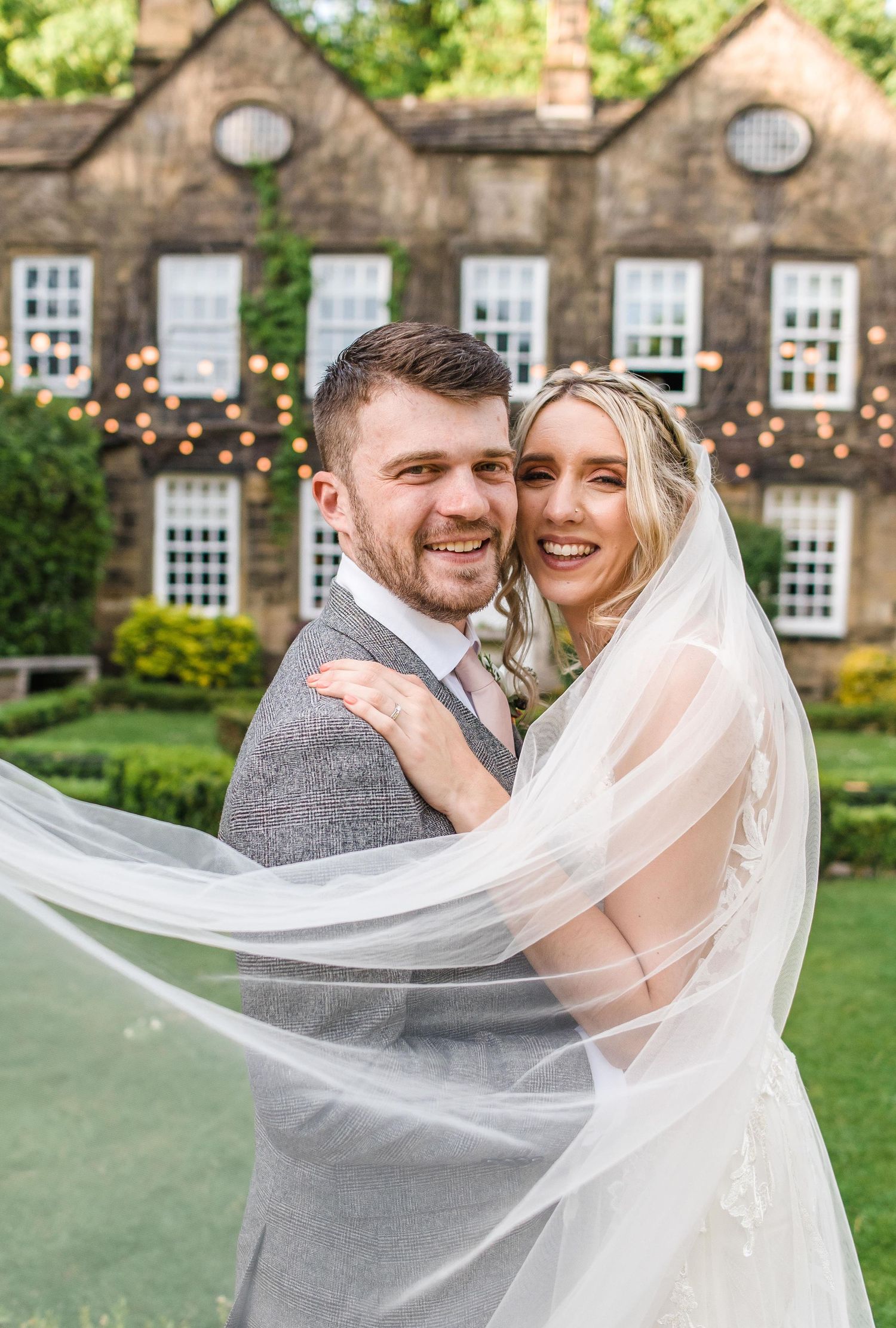 A joyful couple poses in front of a historic stone building with flowering gardens.