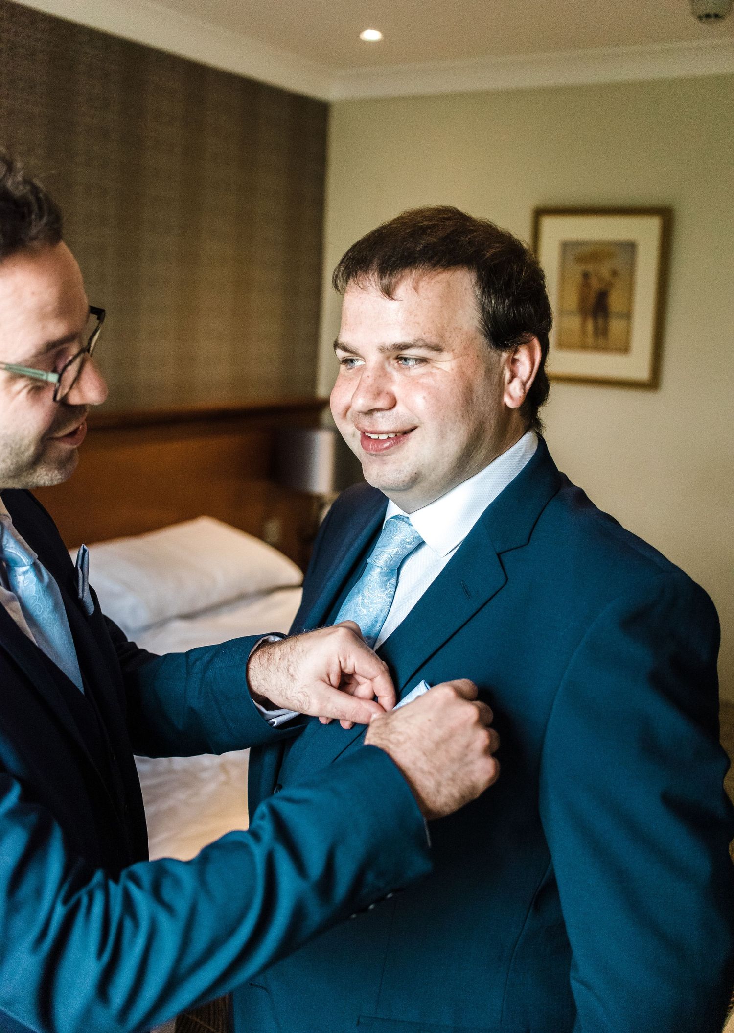 A tender moment of suit adjustment during wedding preparations in a hotel room.