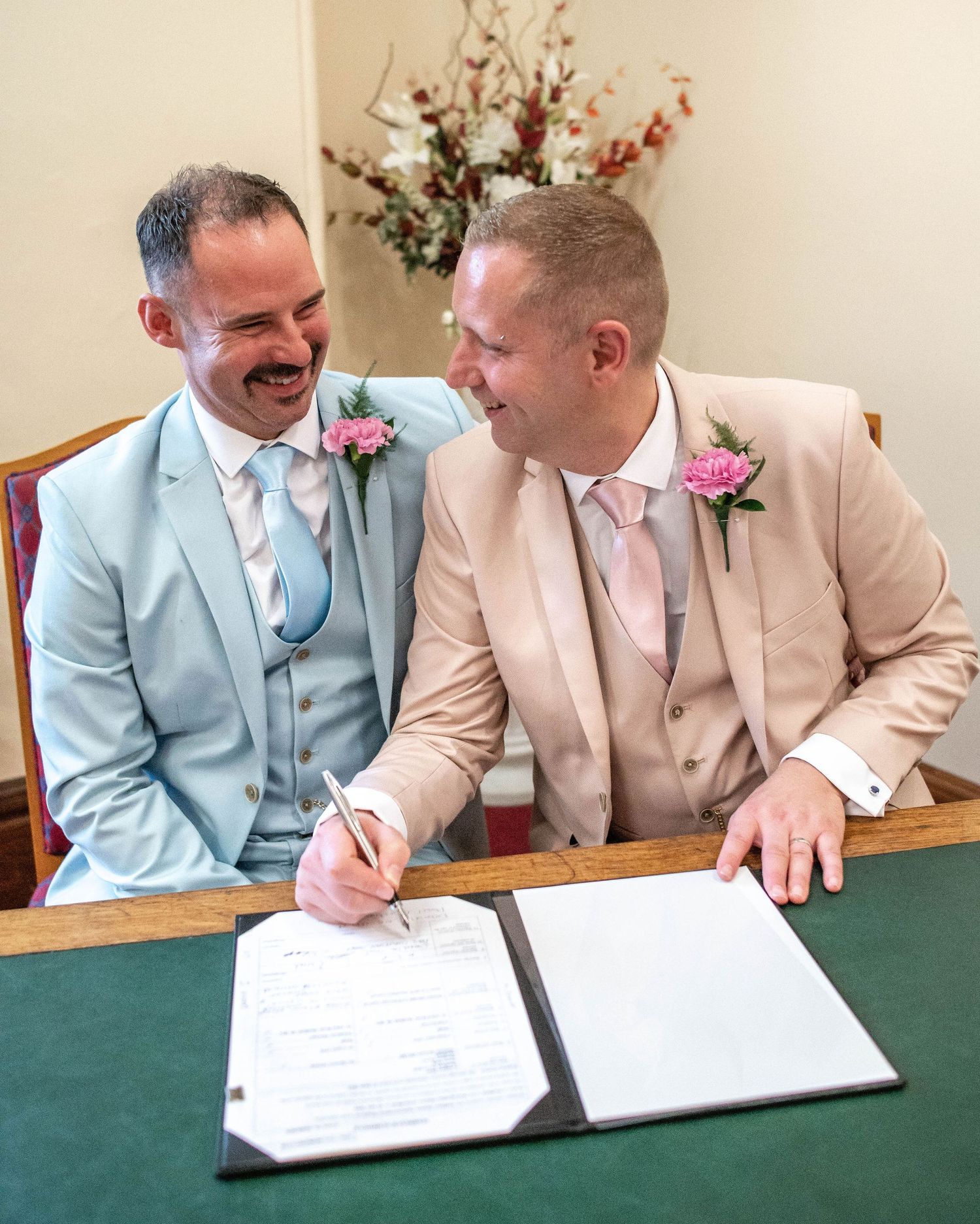 Couple in contrasting suits share intimate moment while signing documents at ceremonial table.