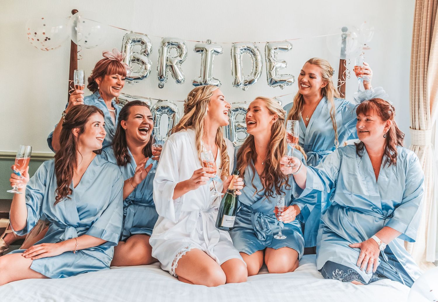 Bridal party in blue robes celebrating on bed with bride in white robe and decorative banner.
