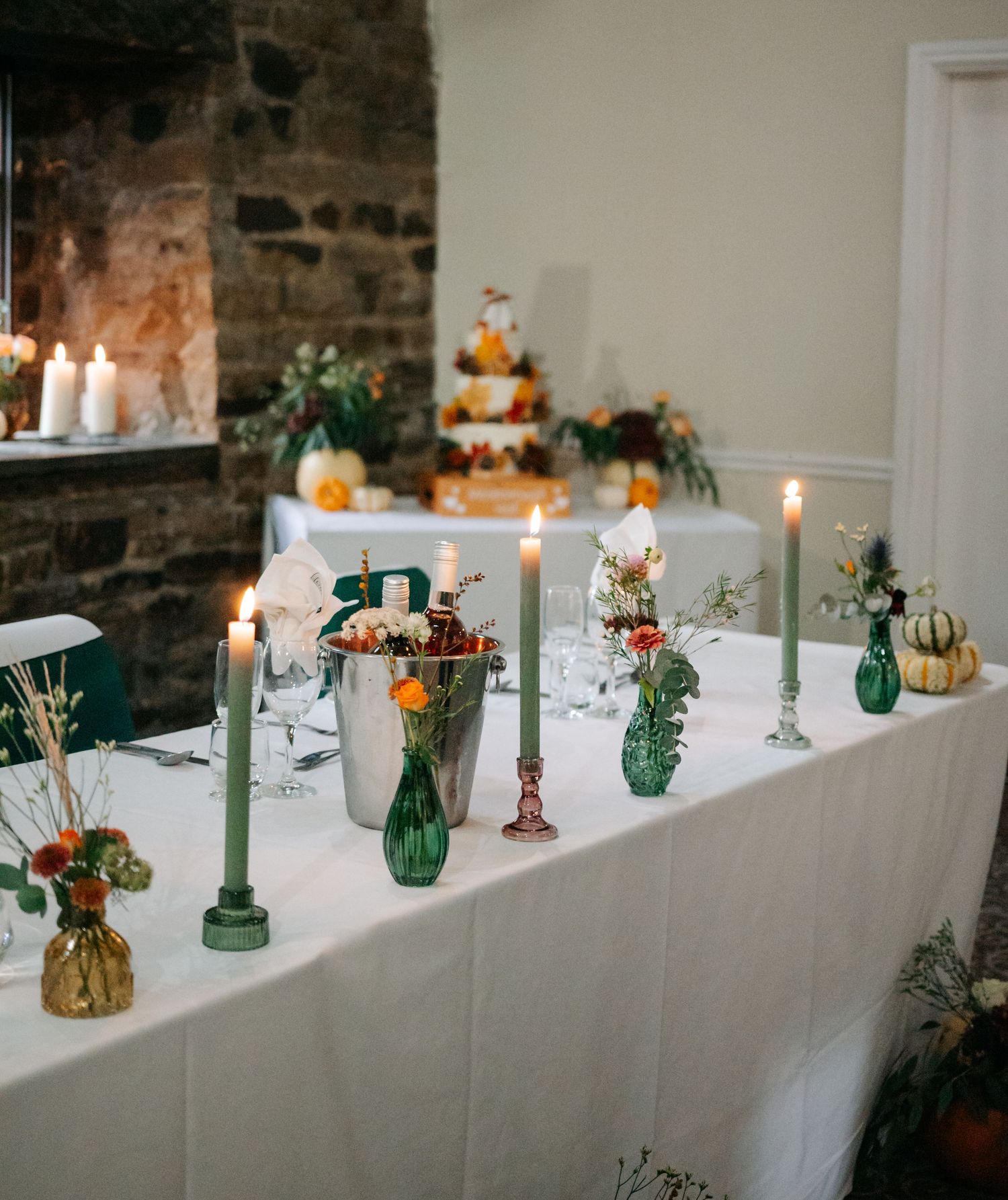 Wedding reception table decorated with green glass bottles and small pumpkins on white tablecloth.