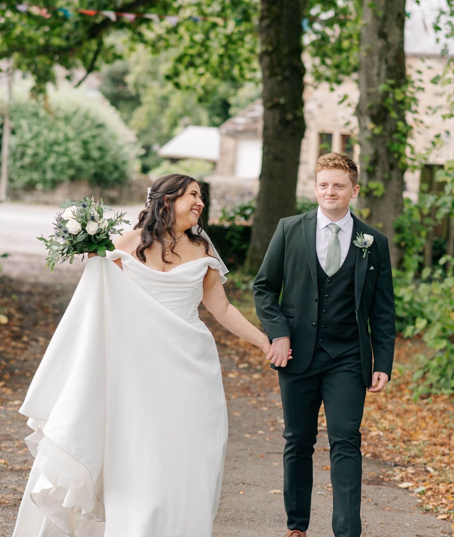 Newlywed couple walking together on a tree-lined path covered in autumn leaves during their wedding day.