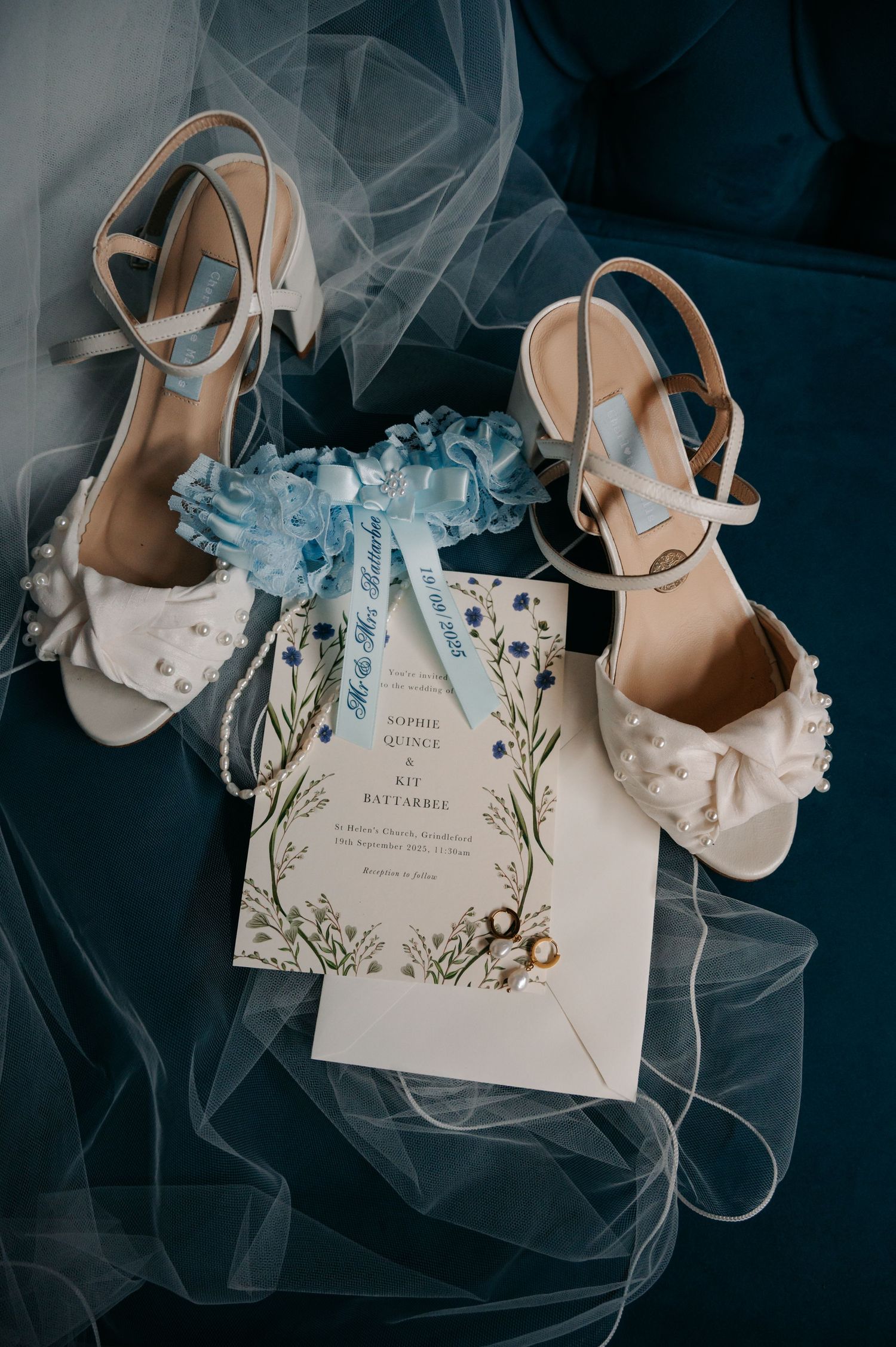 Elegant wedding details including white heels, delicate tulle veil, and floral invitation card on dark background.