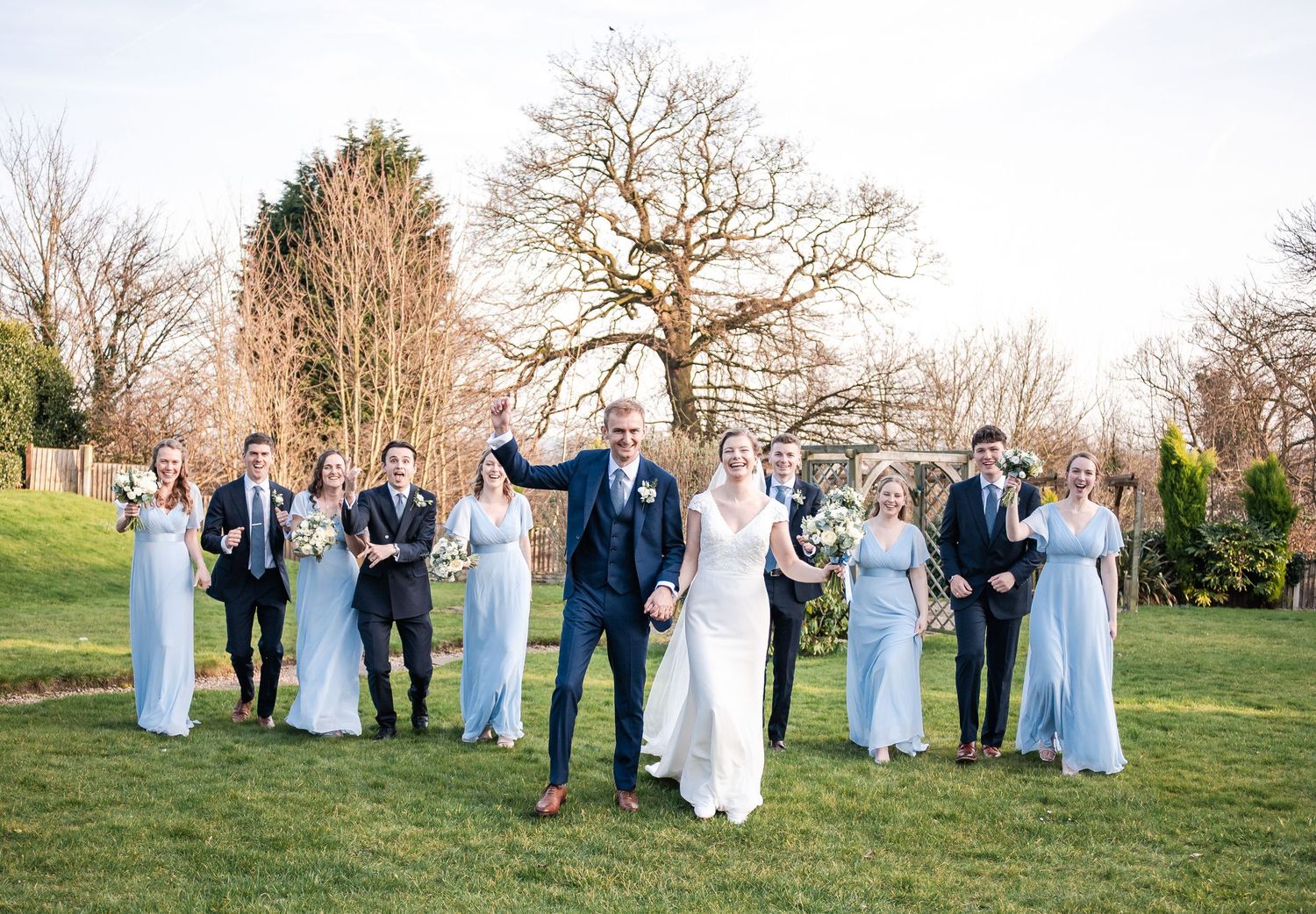 Wedding party poses for photos outdoors in light blue dresses against a natural backdrop.