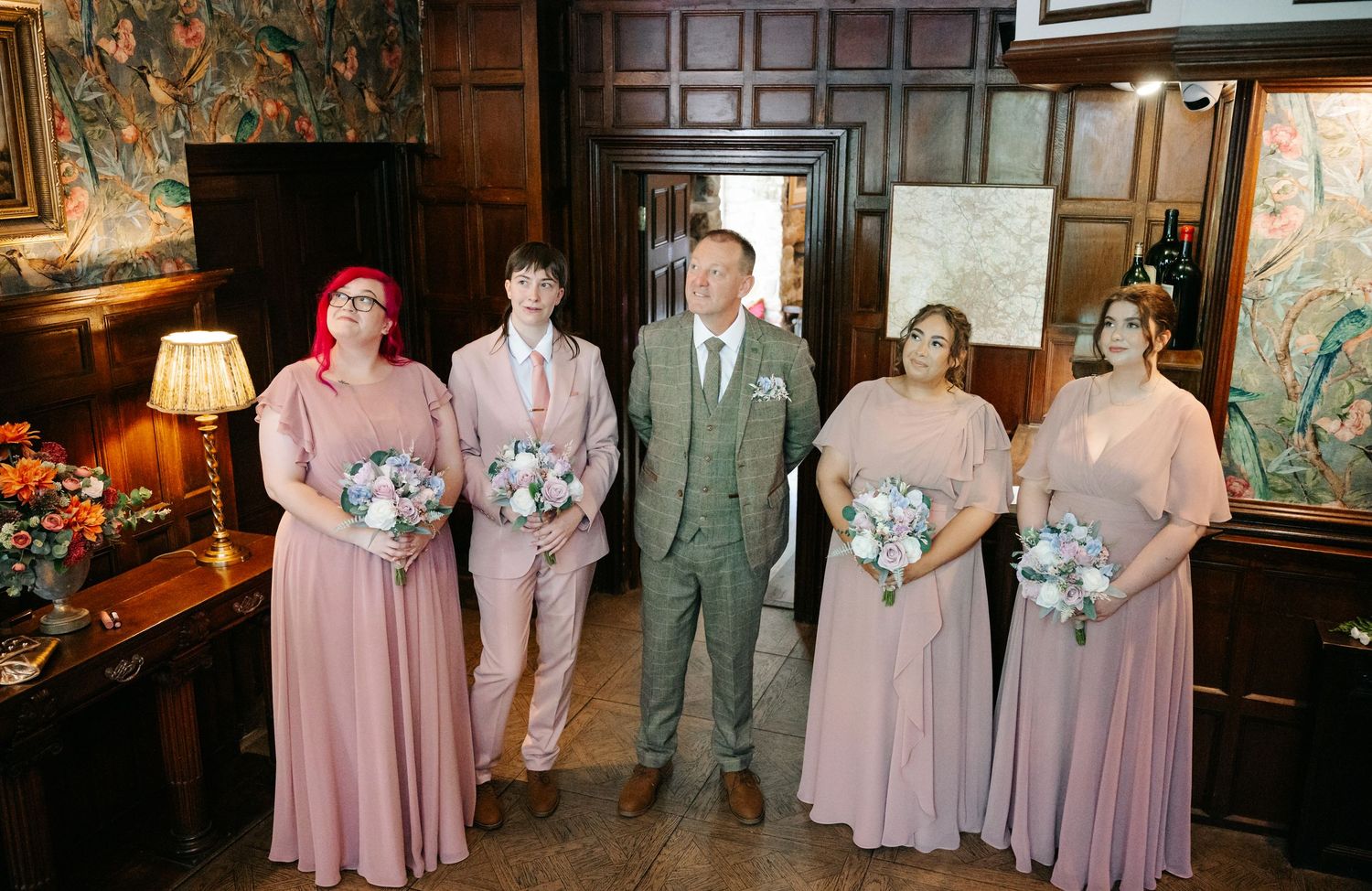 Wedding party posing indoors, groom in grey suit with bridesmaids in dusty pink dresses holding bouquets.