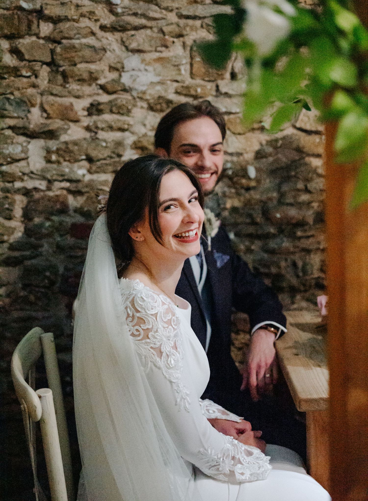 A wedding couple poses together against a rustic stone wall with natural lighting.