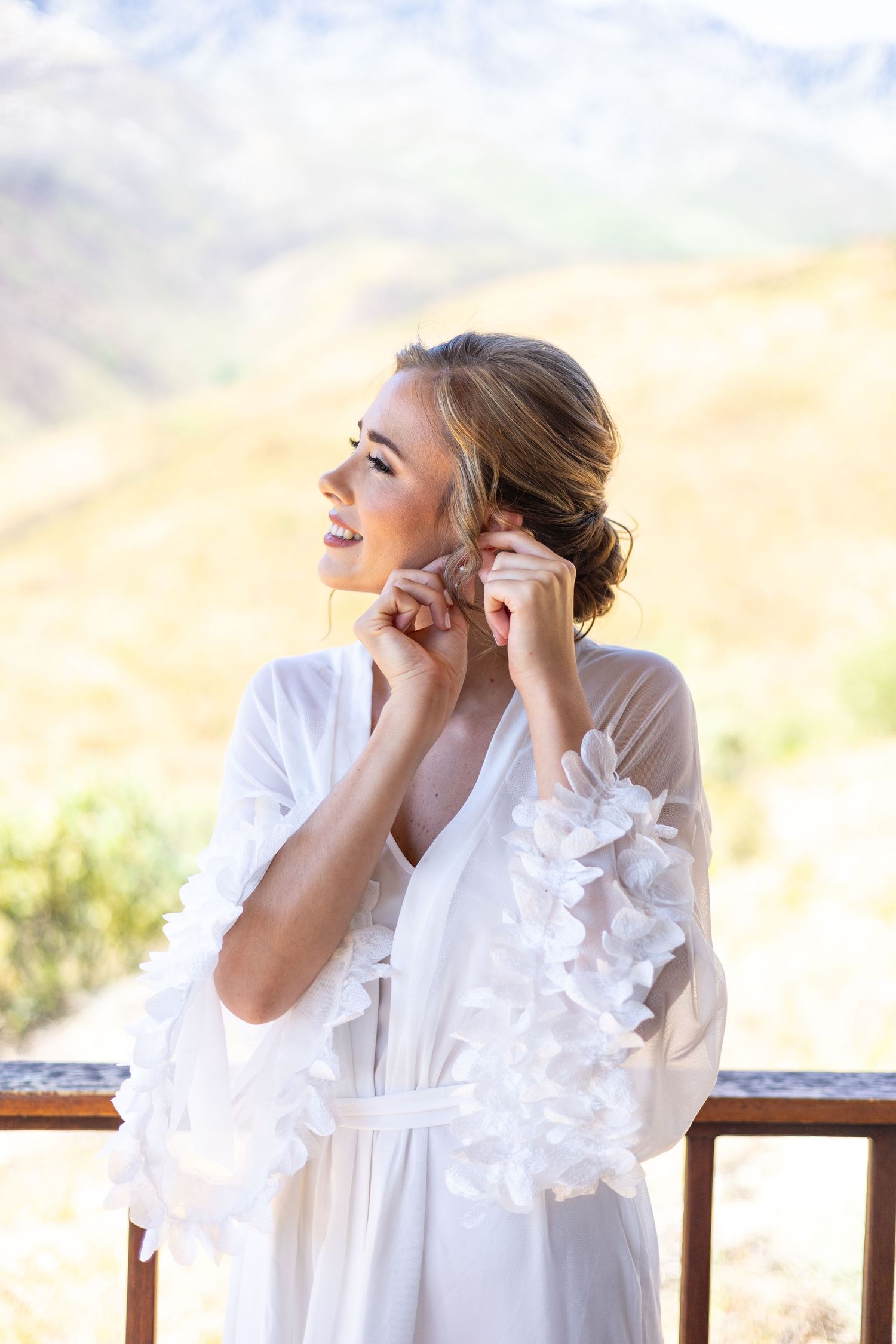 Woman in flowing white dress with ruffled sleeves smiles while putting on earrings on an outdoor balcony at sunset.
