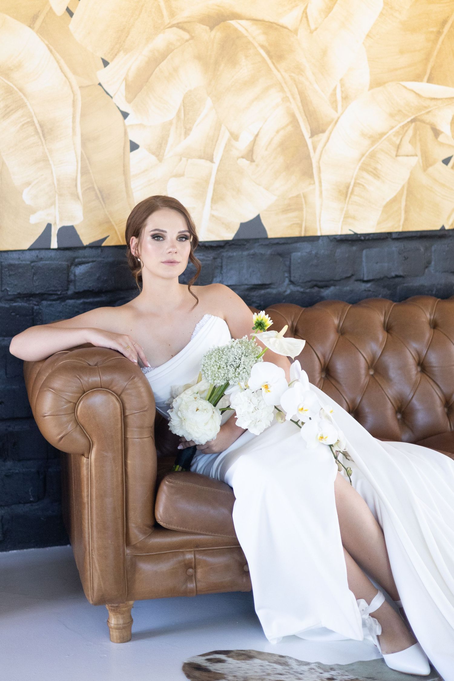 Someone in an elegant white wedding dress reclines on a brown leather Chesterfield sofa holding a white floral bouquet.