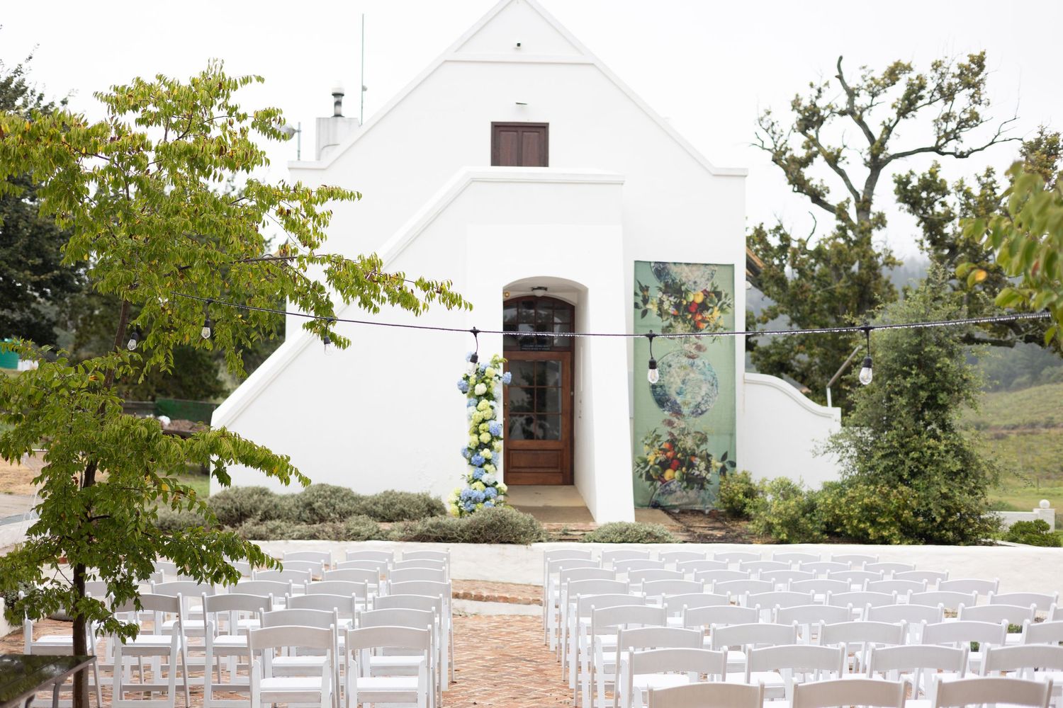White chapel with wooden door and rows of white chairs set up for an outdoor wedding ceremony on stone pathway.