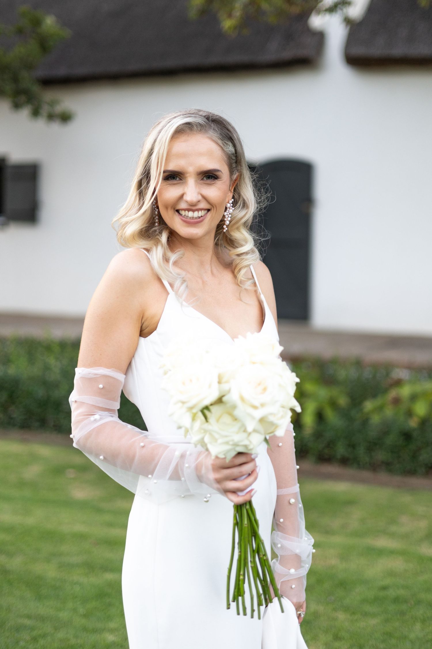 A bride wearing a white wedding dress and sheer lace gloves holds a bouquet of white flowers outdoors.