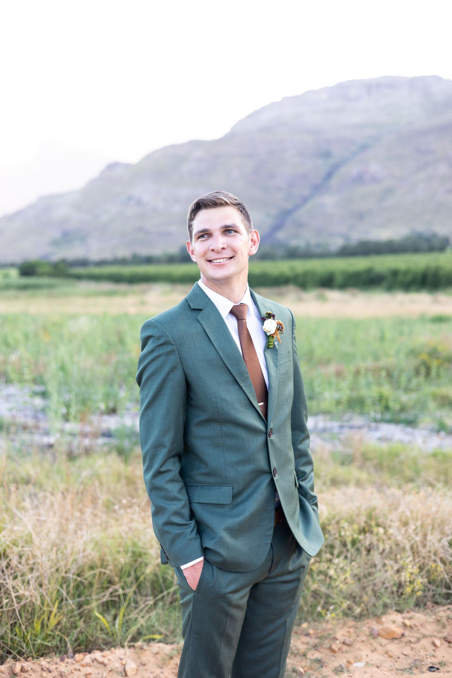 A person in a dark green suit with brown tie poses outdoors in a meadow with mountains in the background.