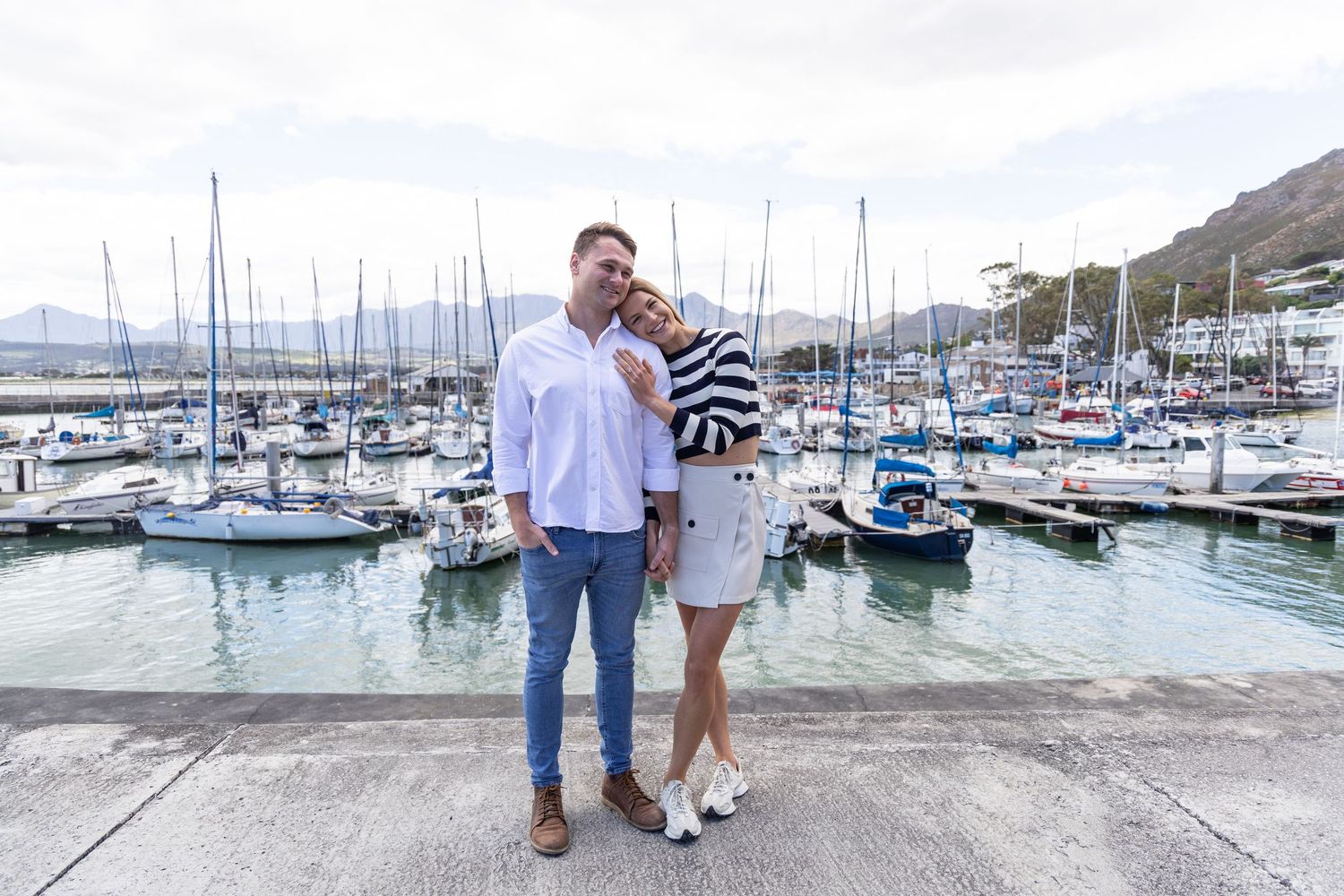 A stylish couple poses together on a dock surrounded by sailboats in a scenic harbor.