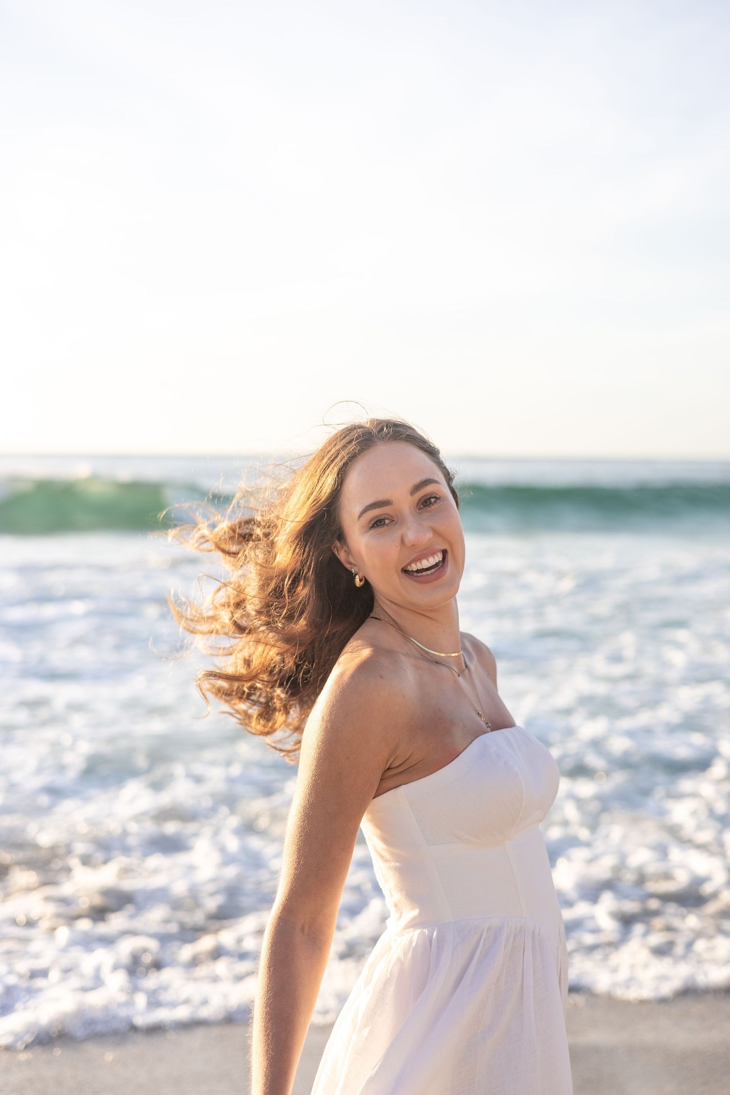 Carefree bride in white dress laughing on beach with waves in background.