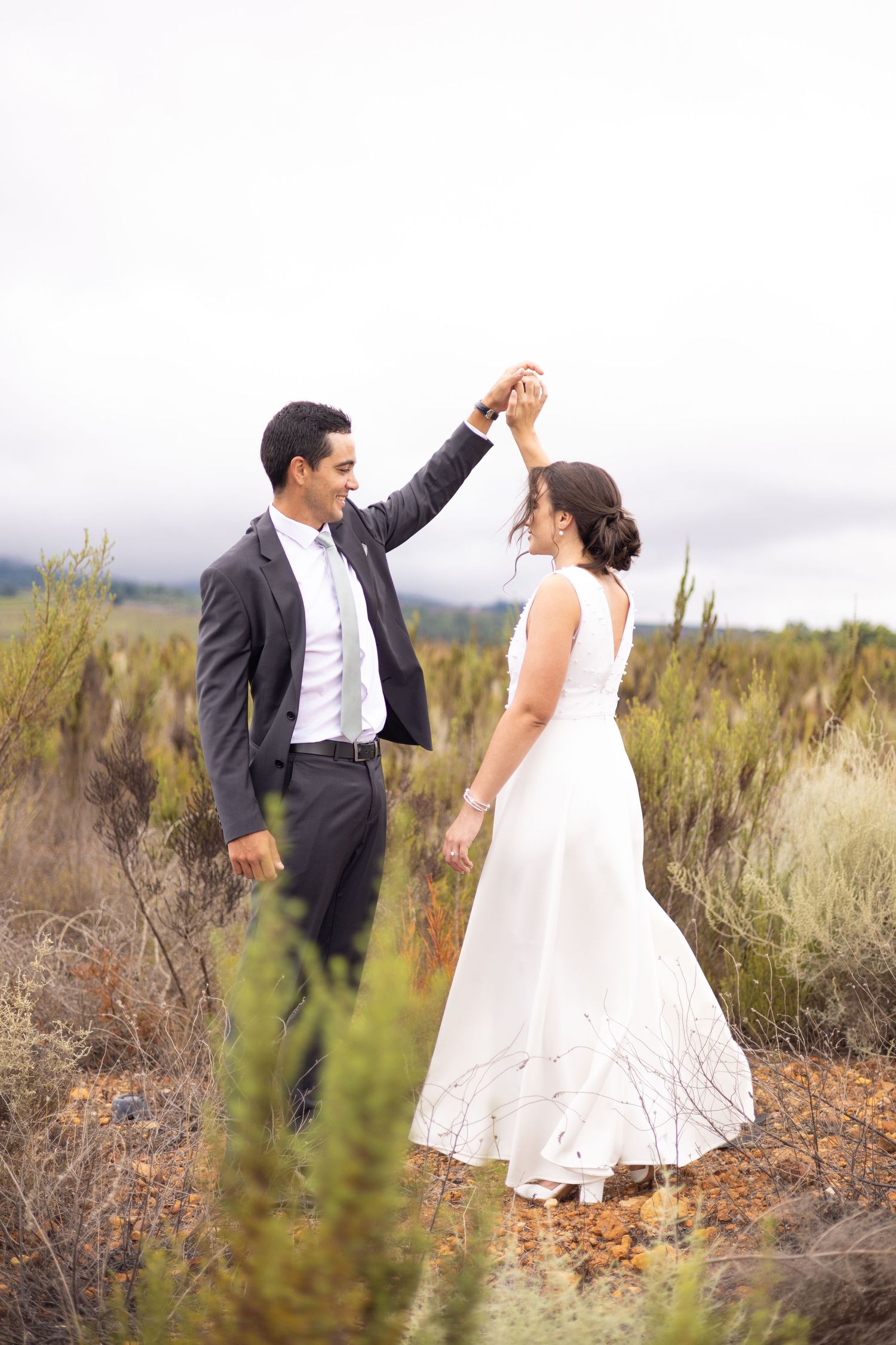 A couple in wedding attire dance together in a desert landscape with sagebrush and cloudy skies.