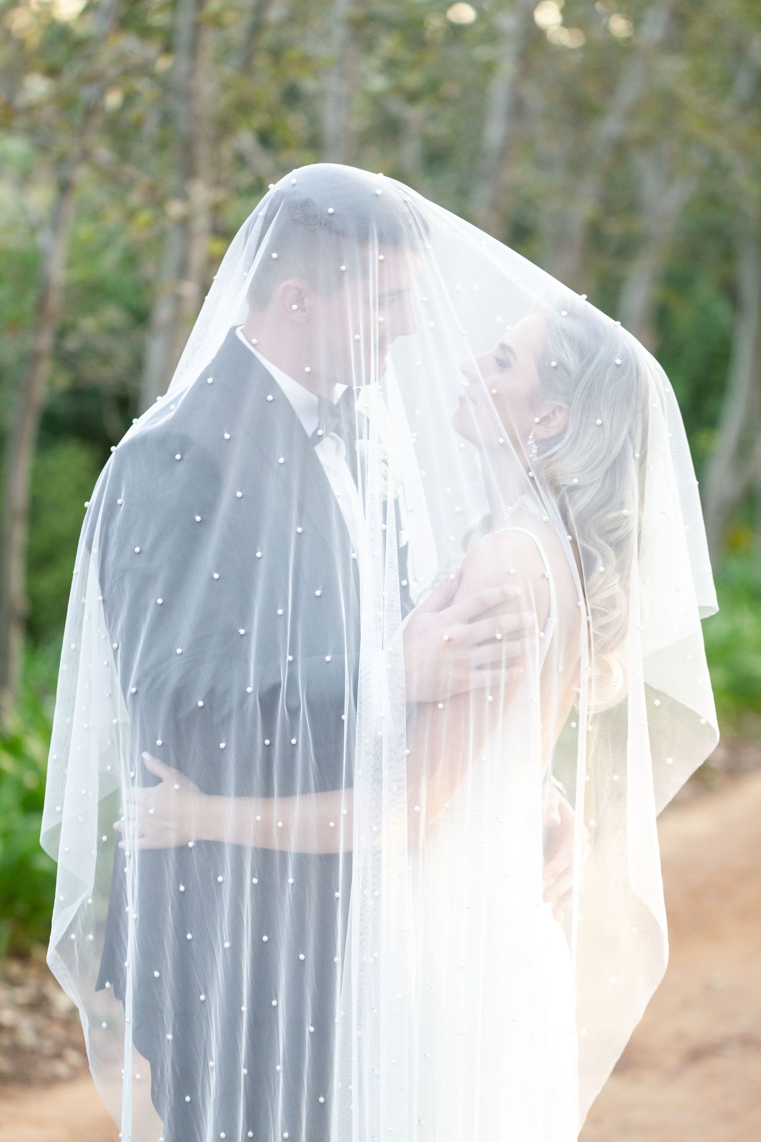 Newlyweds share tender moment under pearl-decorated wedding veil outdoors.