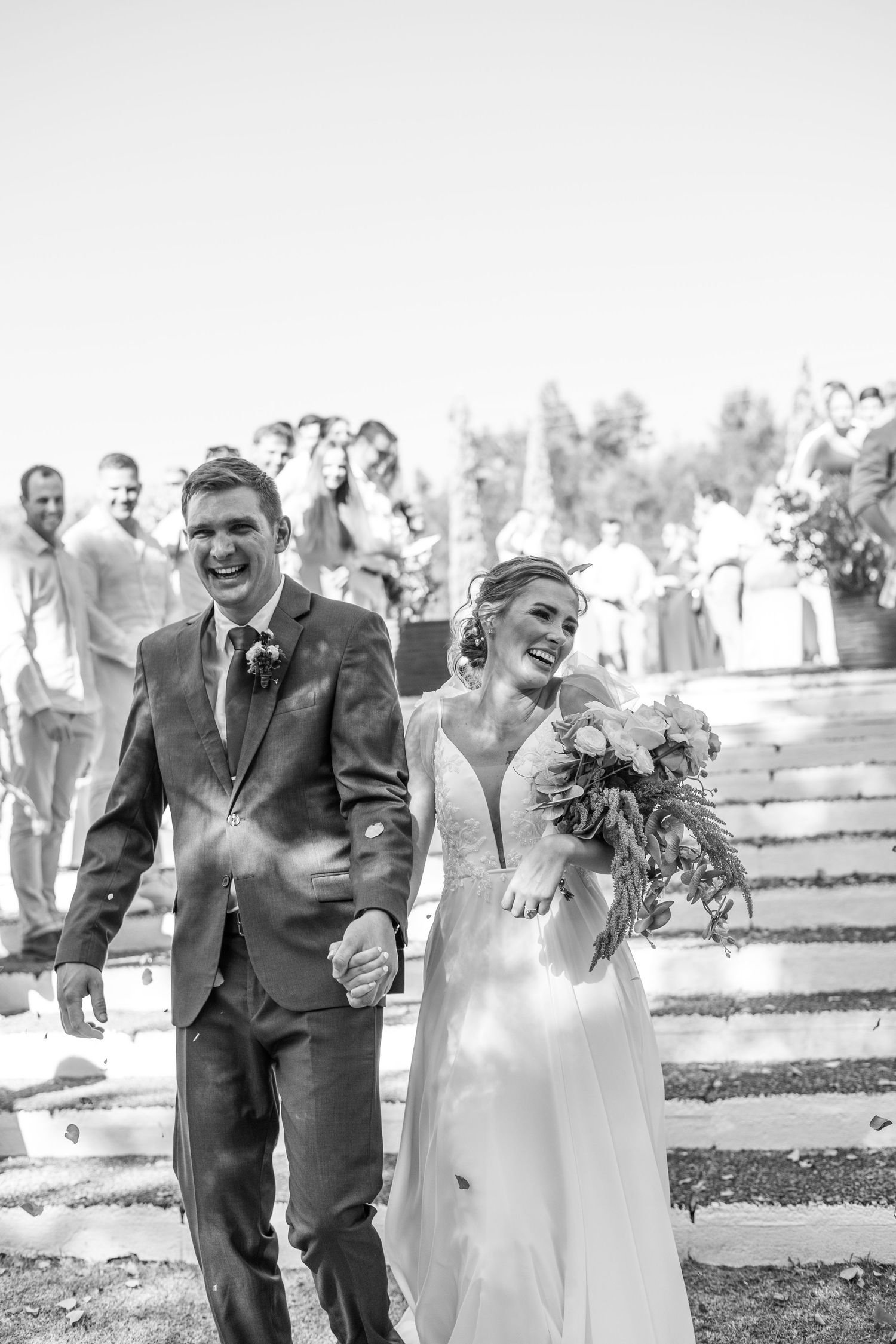 A joyful couple walks down outdoor steps after their wedding ceremony, with guests visible in the background.