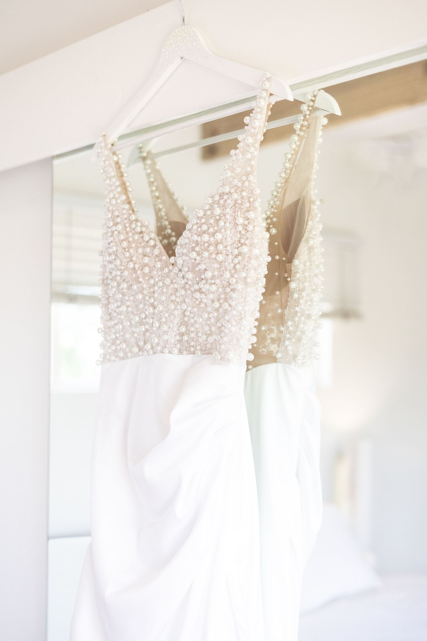 A white wedding dress with delicate pearl beaded bodice hangs elegantly against a light wall.