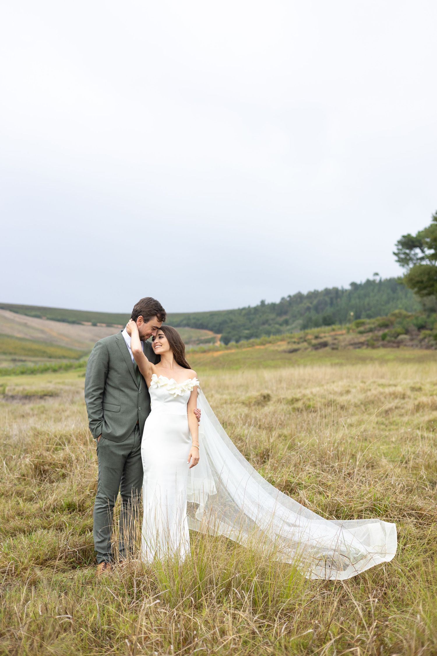 A bride and groom share intimate moments in a scenic meadow with flowing white veil and rolling hills behind them.