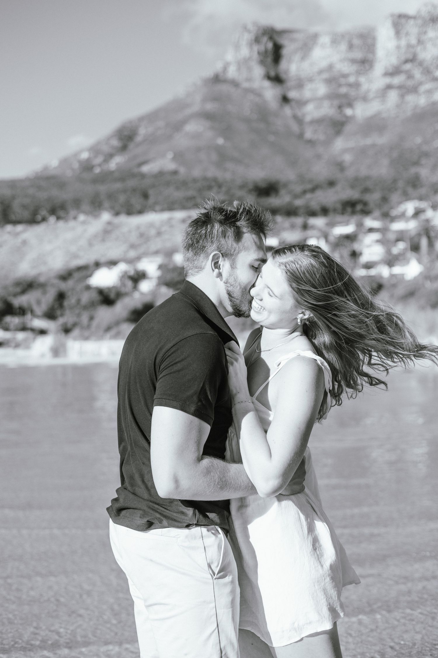 Romantic black and white photo of couple embracing on beach with mountain backdrop.