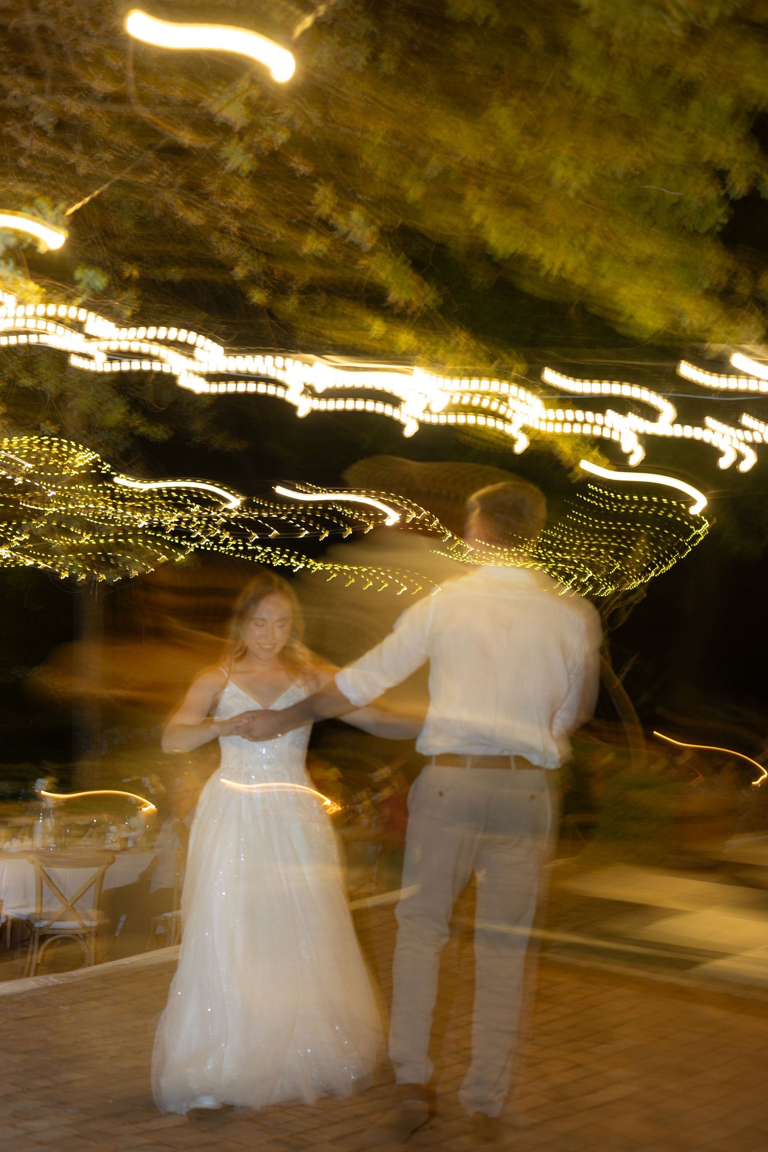 Motion blur captures a romantic dance beneath twinkly string lights at an outdoor evening celebration.