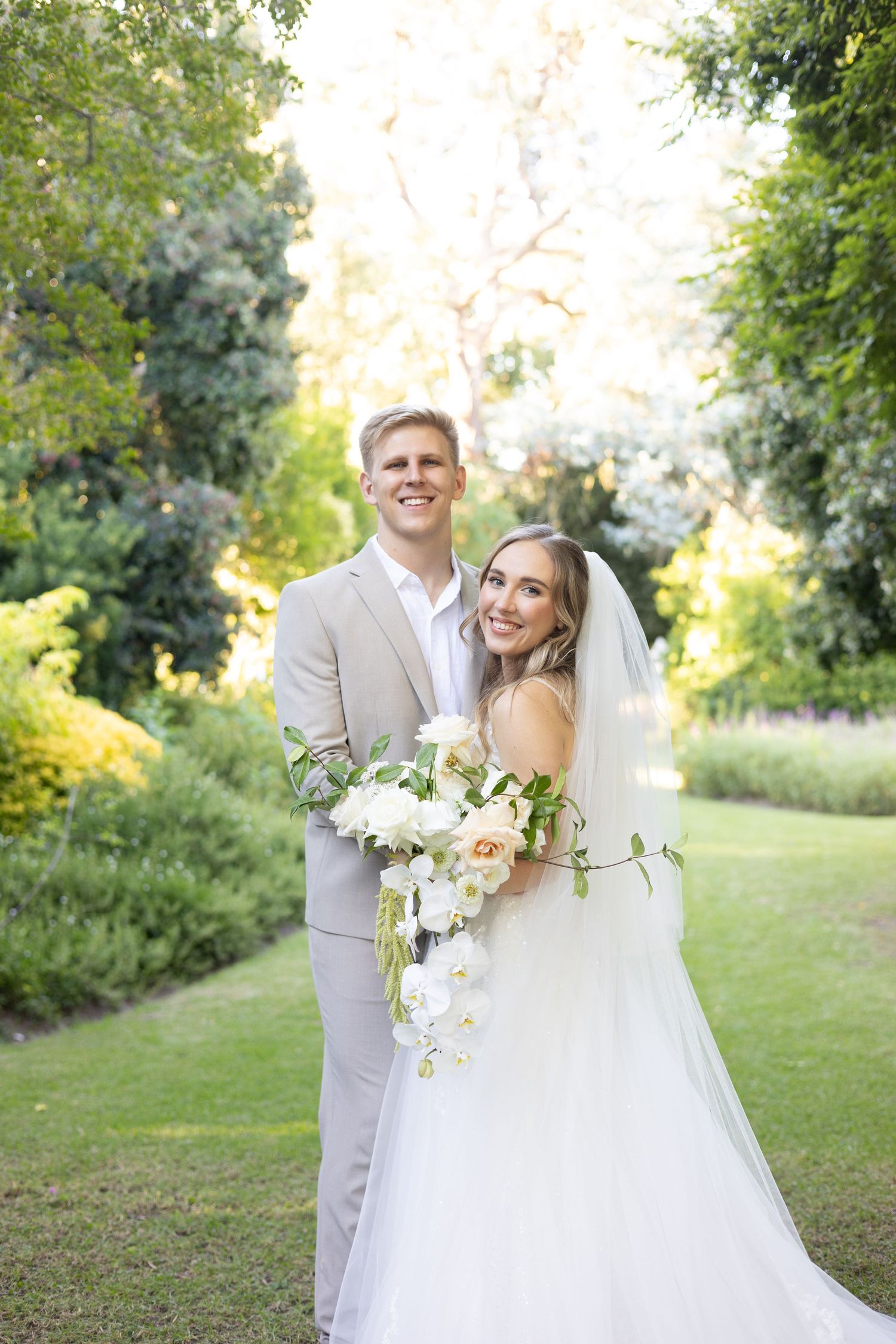 A bride in a white gown and groom in light gray suit pose together in a lush garden setting with white floral bouquet.