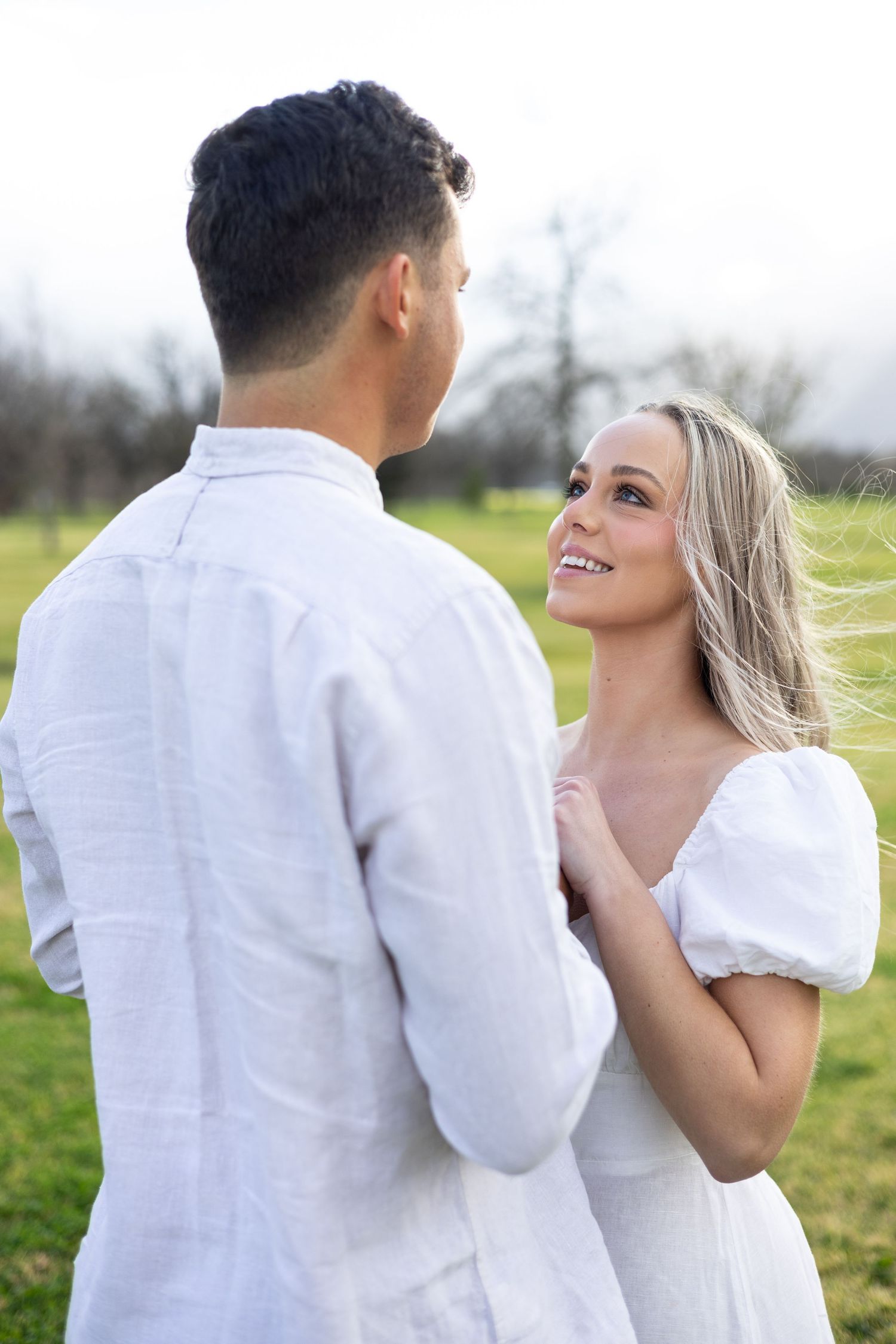 A romantic couple in white outfits sharing an intimate moment while standing in a grassy field on a cloudy day.