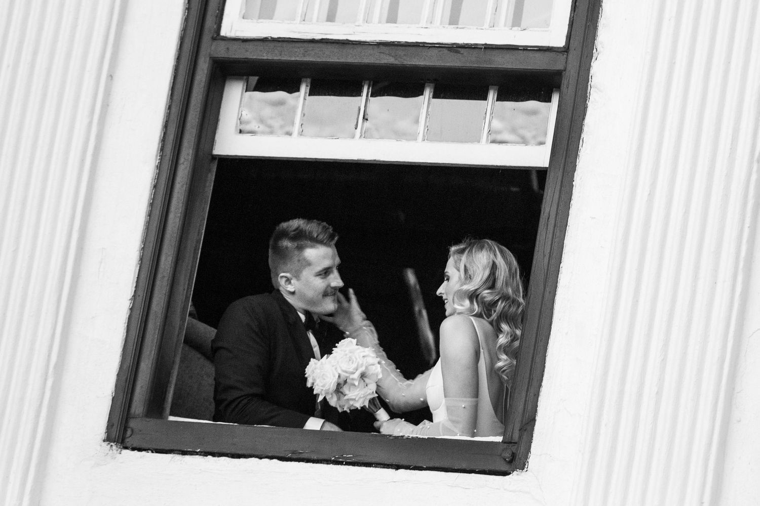 Wedding couple sharing playful moment through rustic window frame.