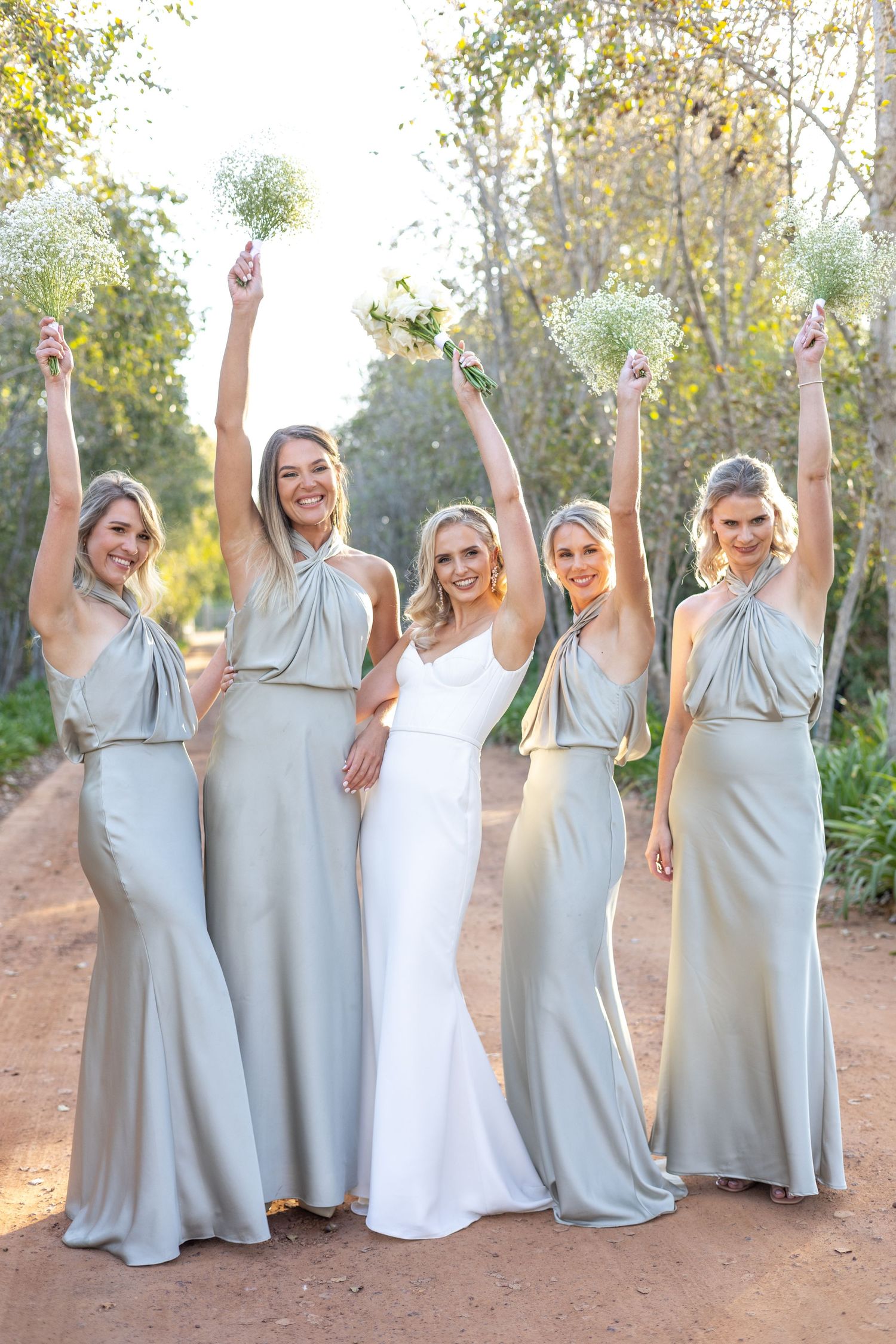Bridal party in pale gray dresses pose with bouquets on rustic path.