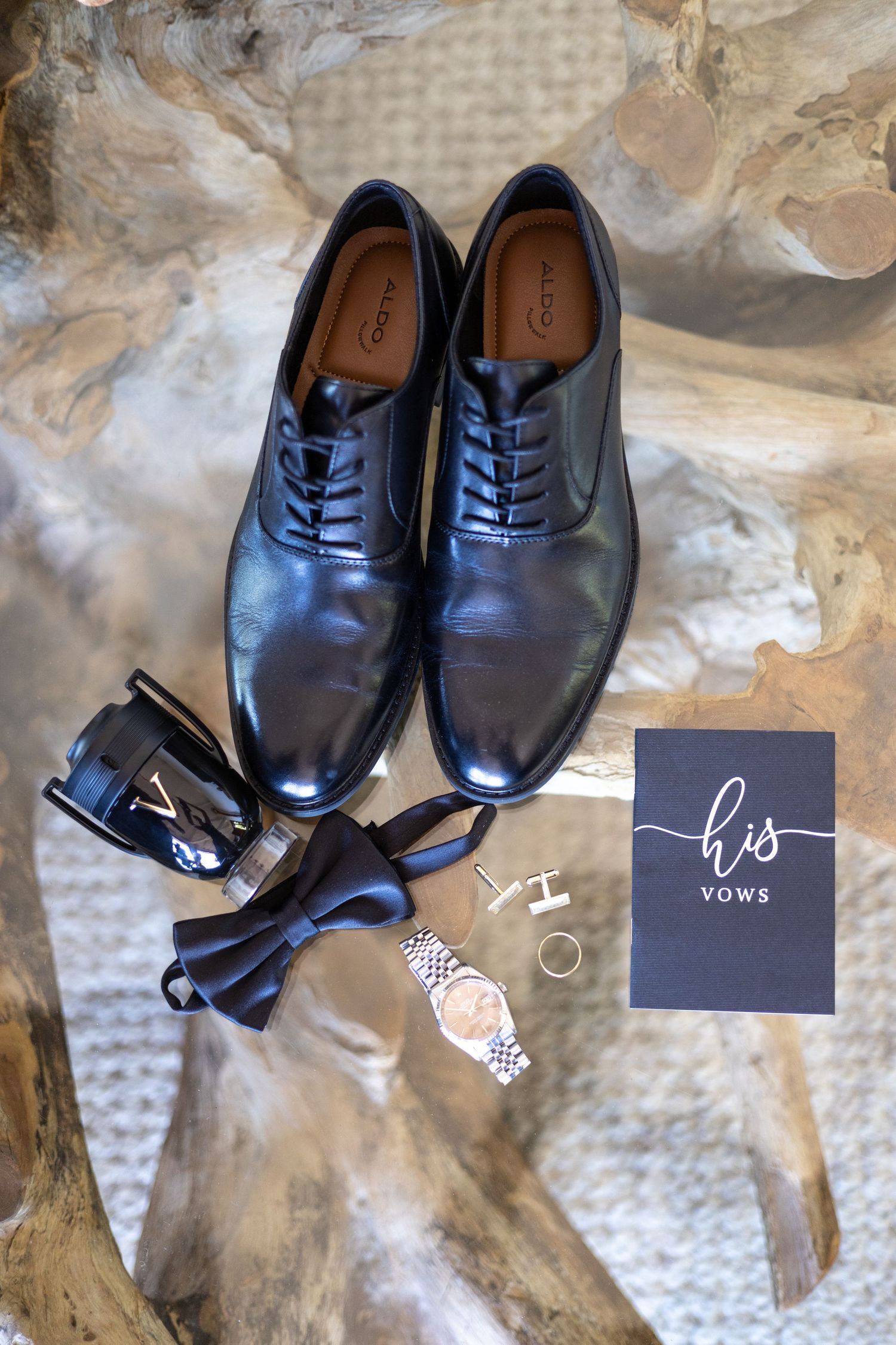 Wedding accessories including black dress shoes, bow tie, watch and vows book arranged on textured fabric.