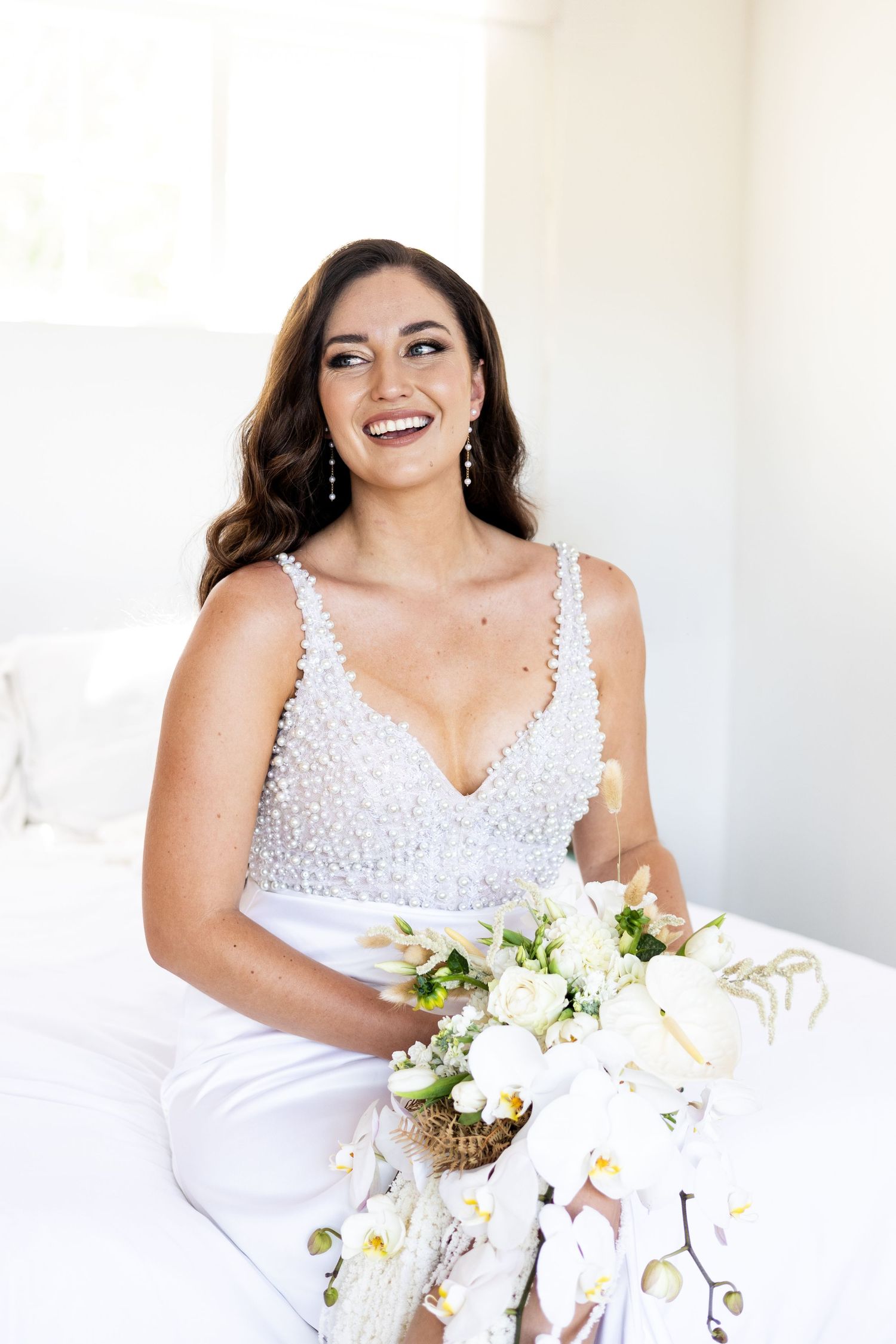 A bride wearing a beaded white wedding gown holds a white and green floral bouquet while smiling and sitting.