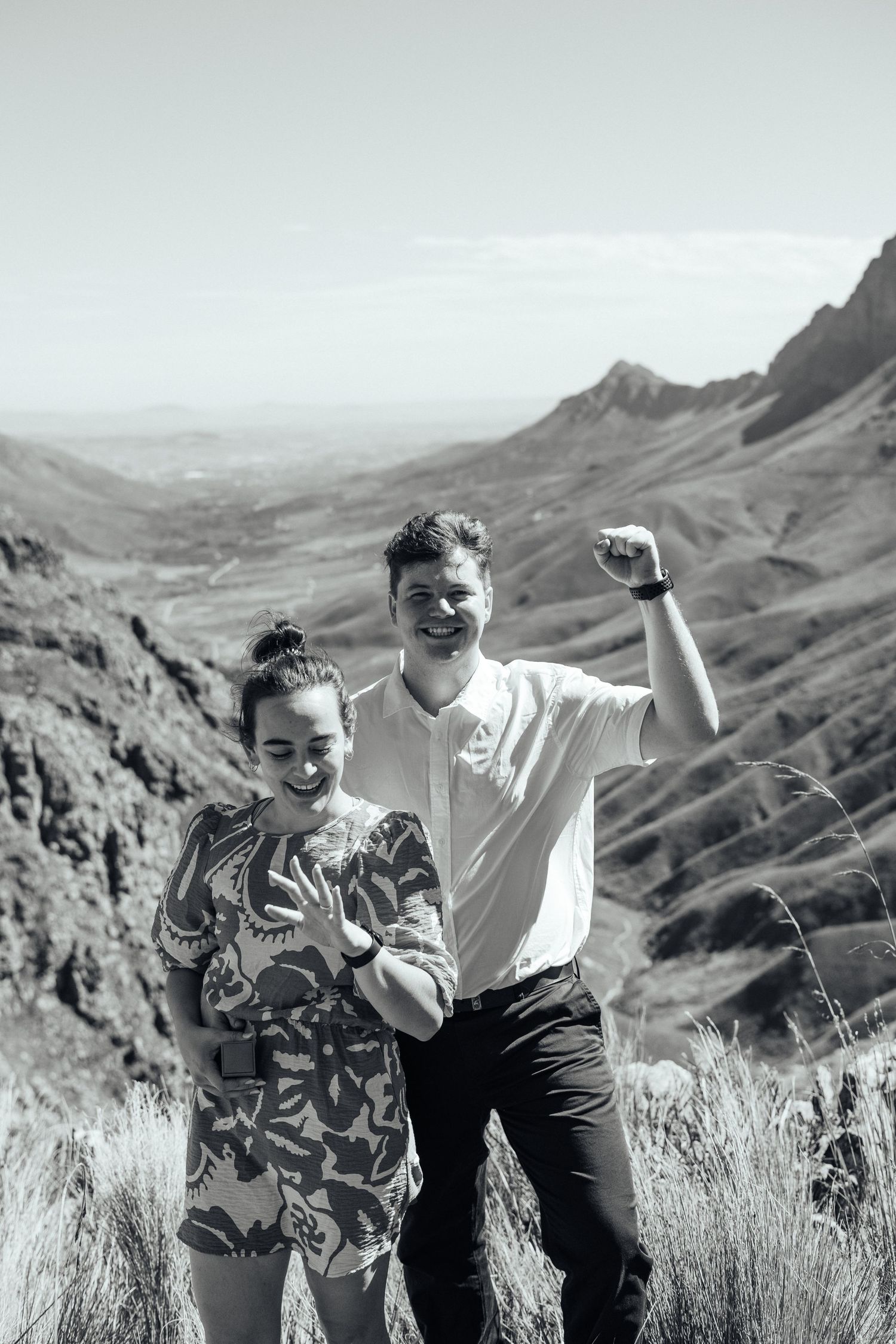 A joyful couple celebrates their engagement with the dramatic mountain landscape behind them.