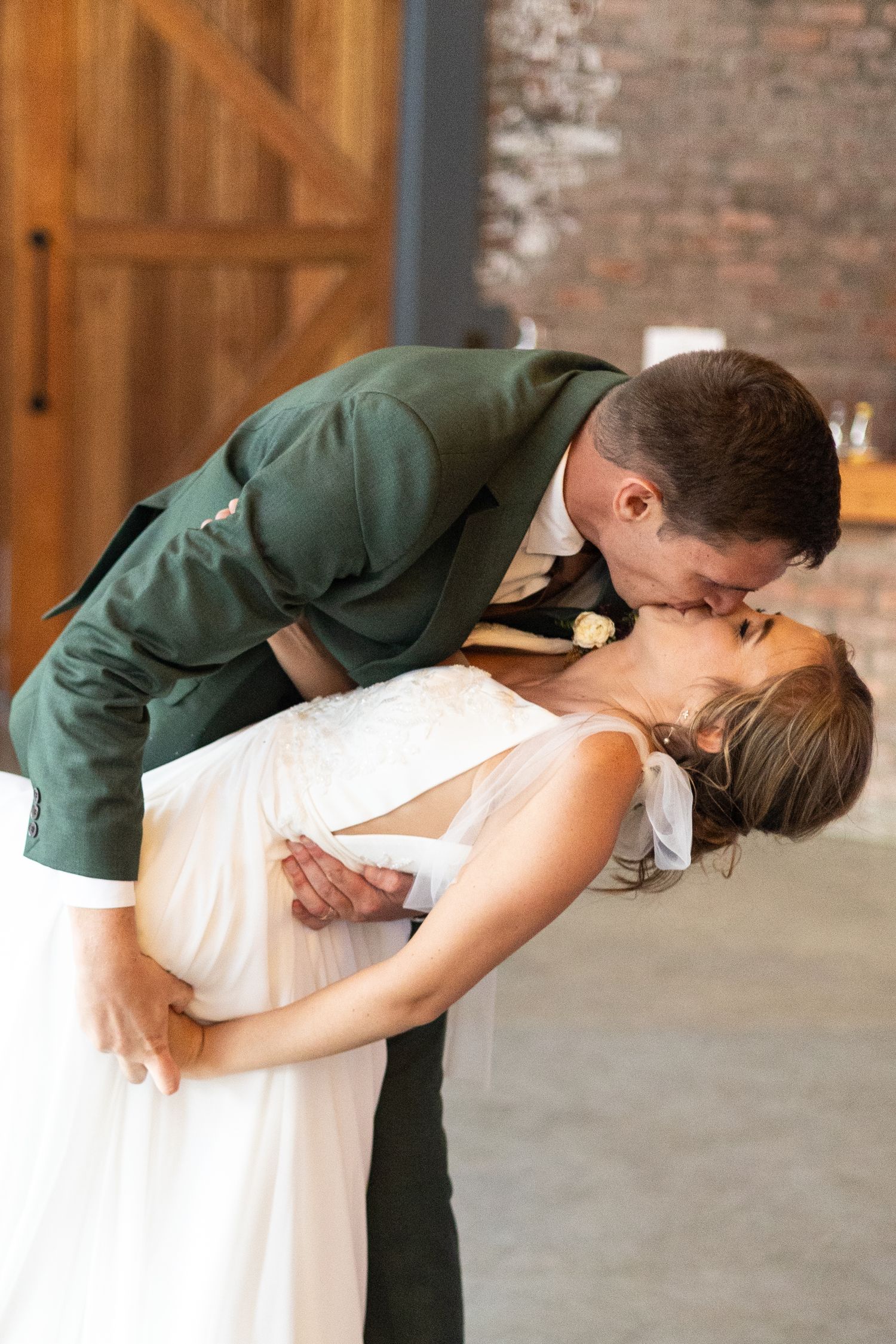 A romantic moment at a wedding ceremony as a bride in white dress dips backward during a kiss in a rustic venue.