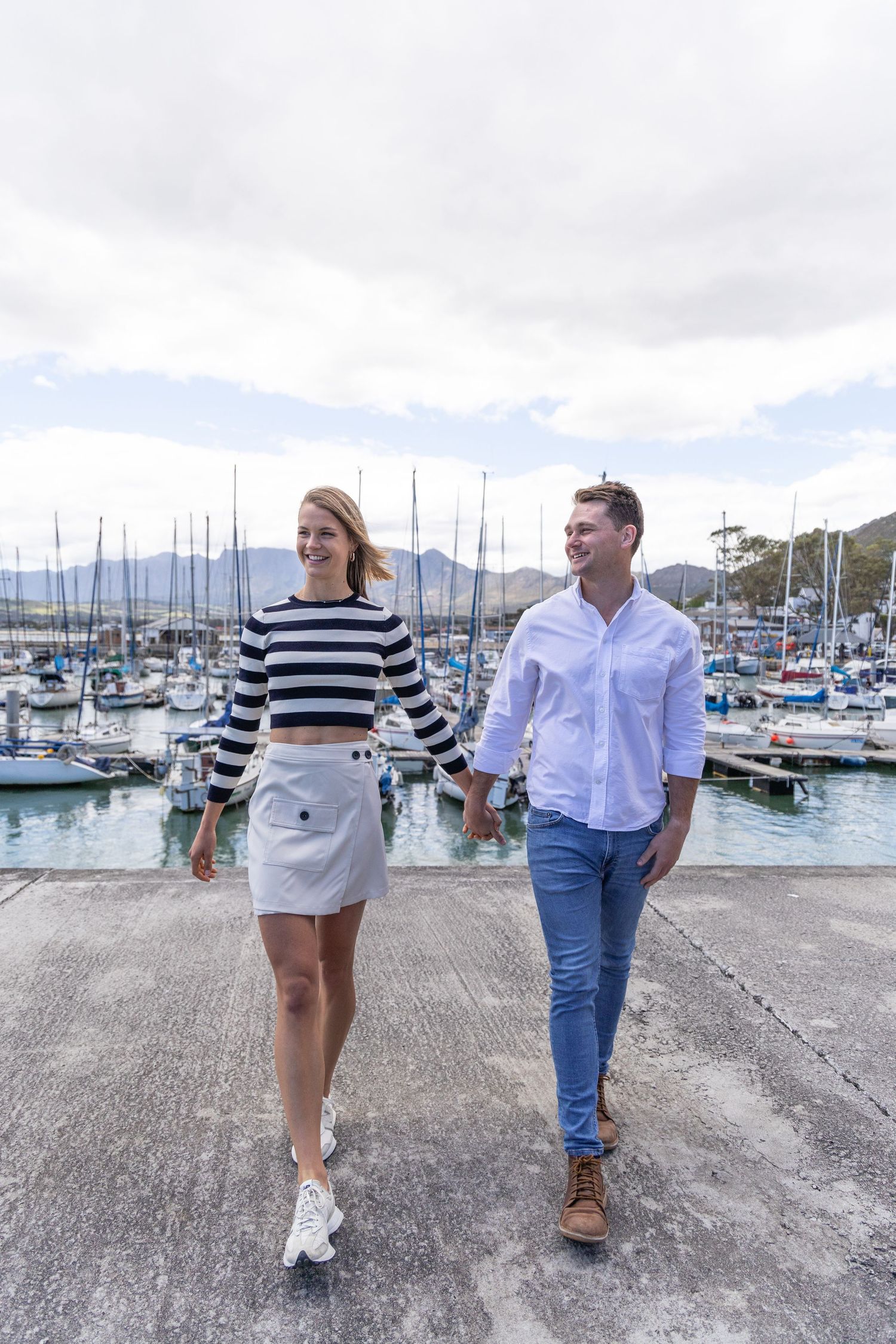 A couple takes a romantic stroll along a marina walkway with boats and mountains in the background.
