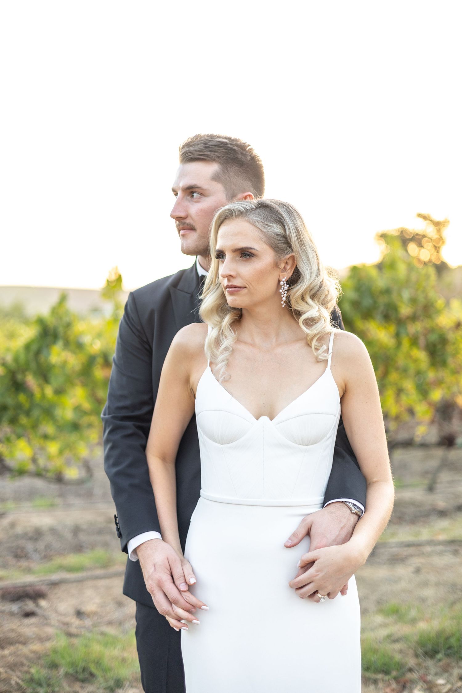 Romantic wedding couple pose together outdoors during golden hour against rustic natural backdrop.
