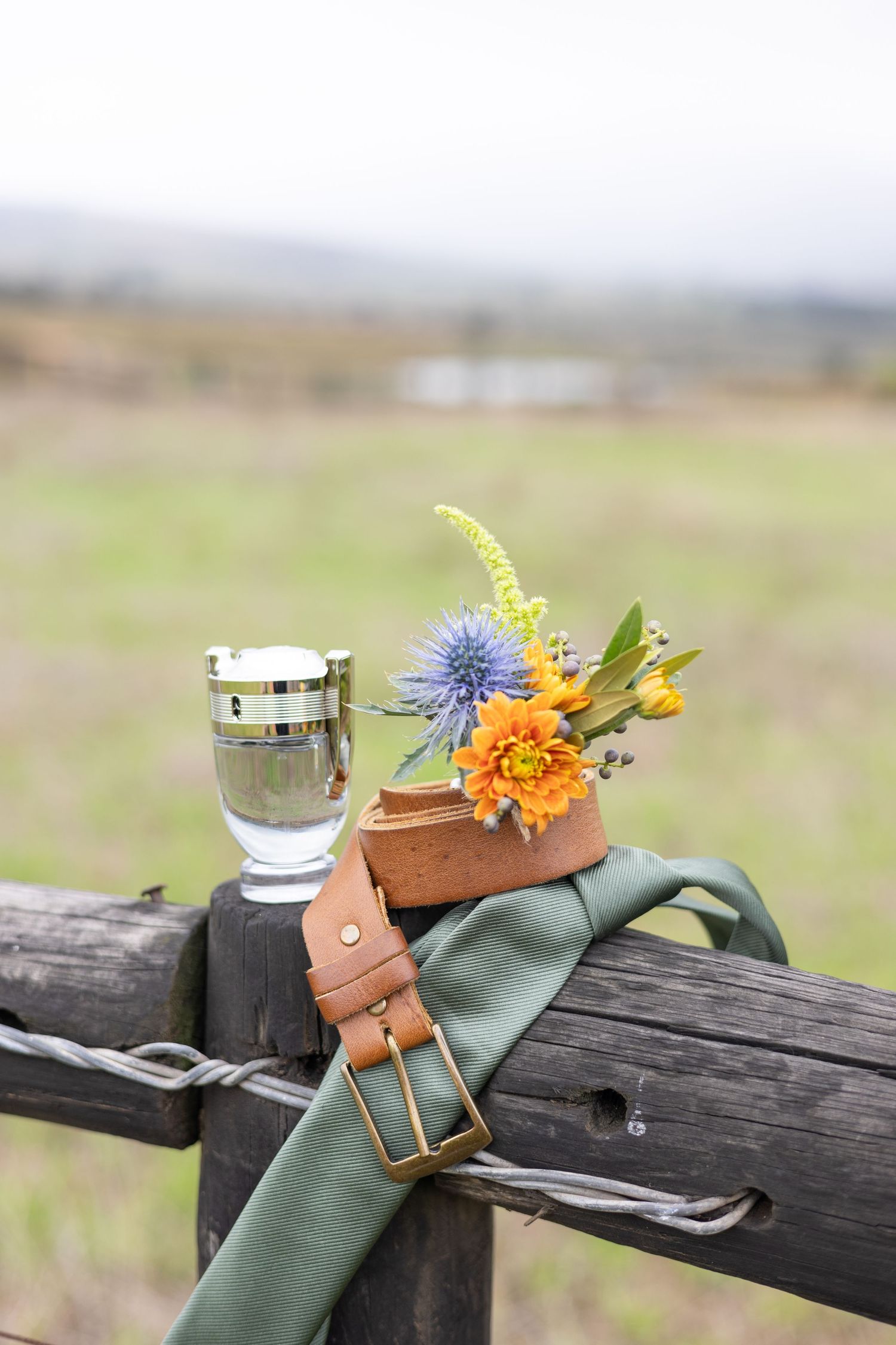 Rustic outdoor setting with a leather belt and colorful wildflowers arranged on a wooden fence with a glass of water.
