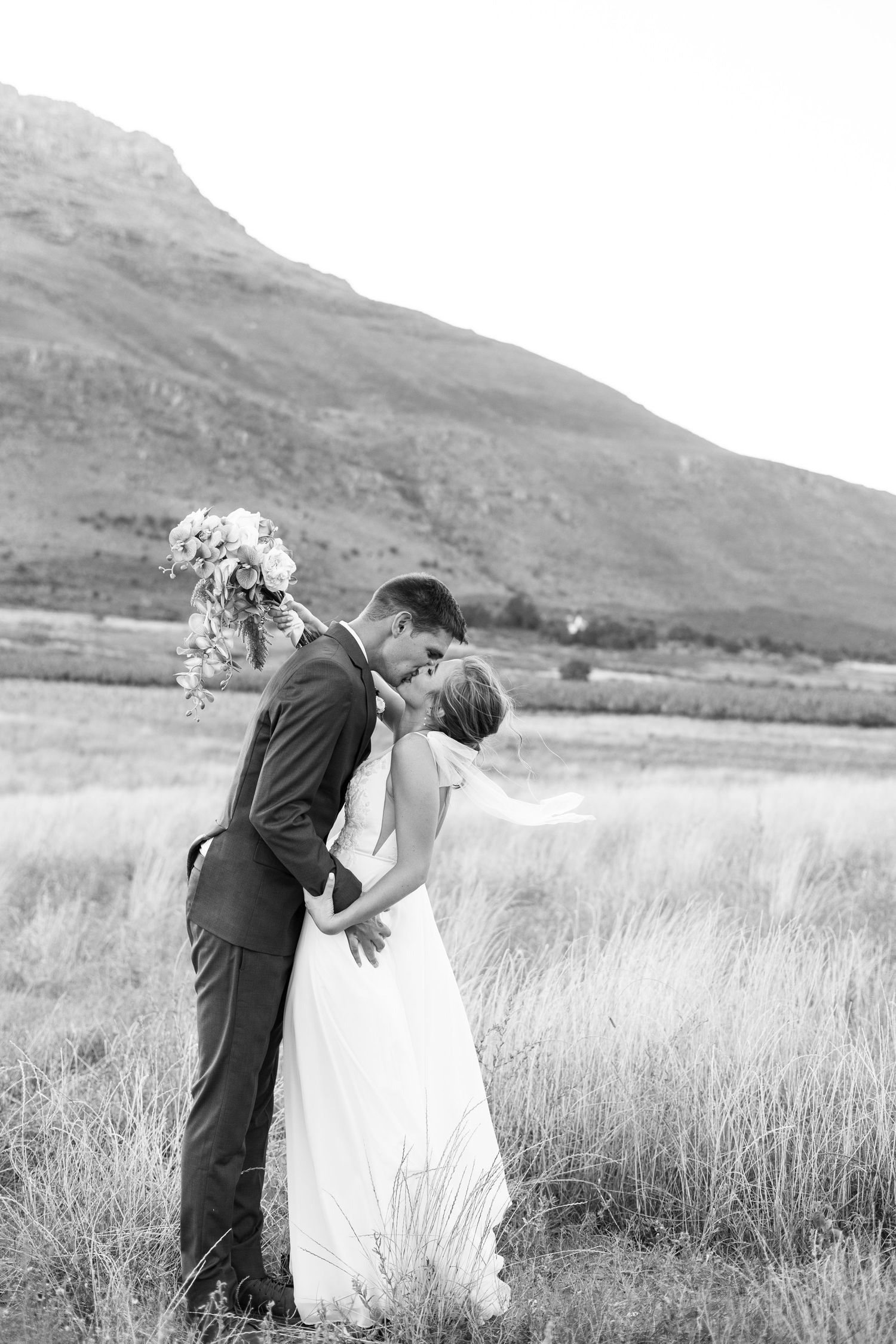 A romantic black and white wedding portrait of a couple embracing in a field with mountains in the background.