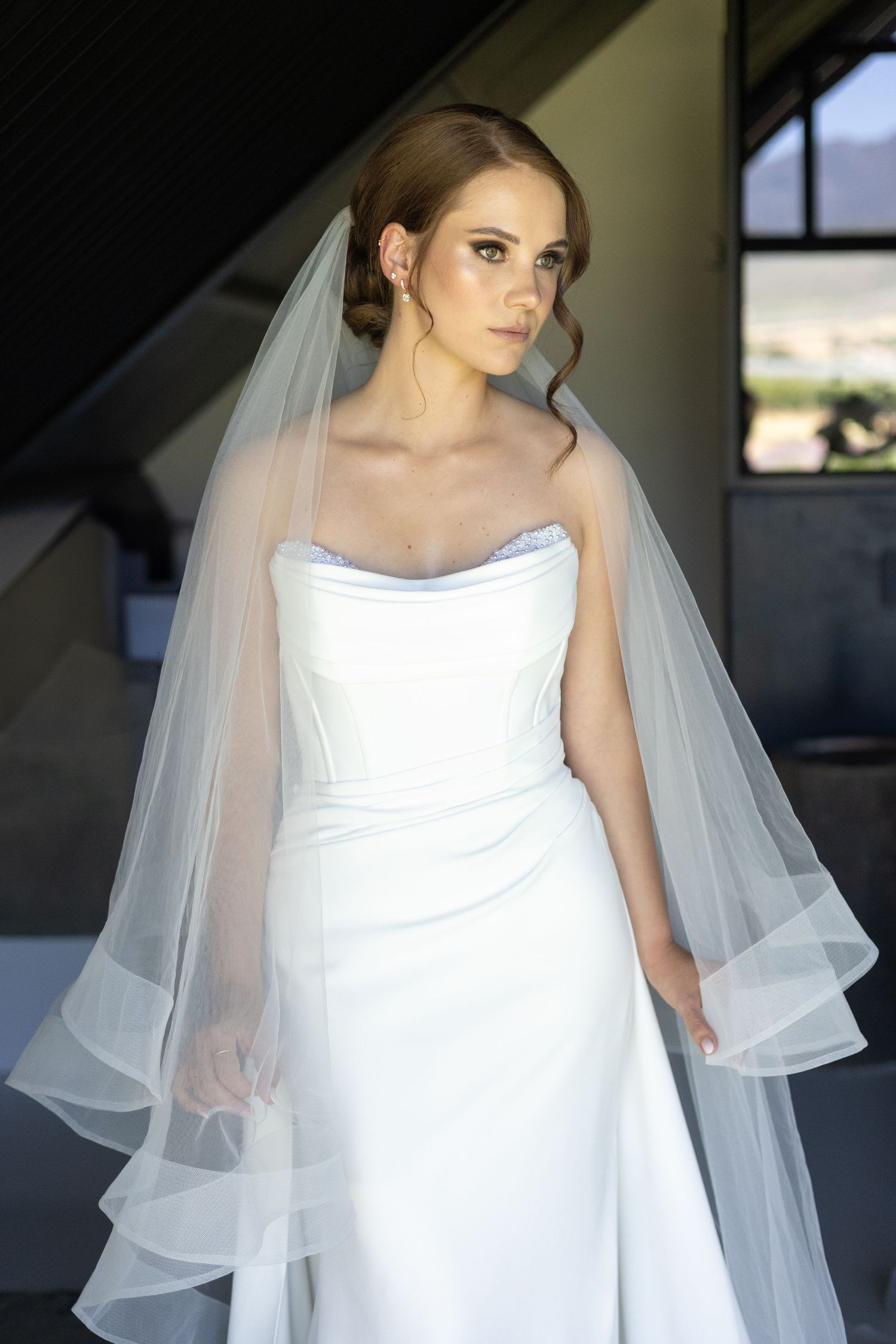 Elegant bride wearing a strapless white wedding gown with a flowing tulle veil stands in natural window light.