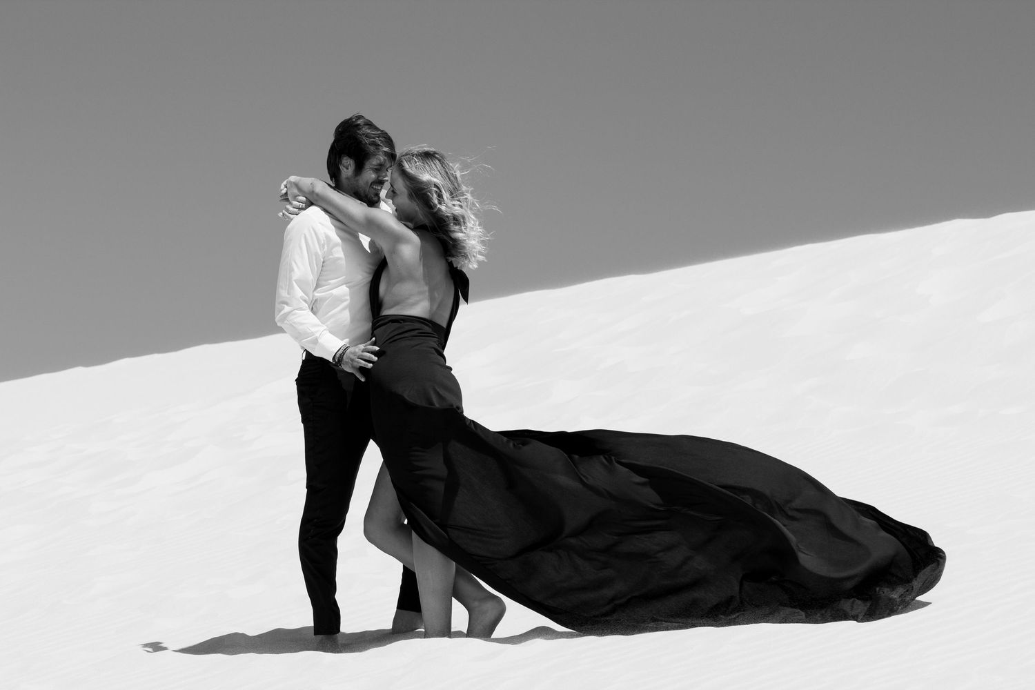 Couple in formal attire poses dramatically on white sand dunes in black and white.