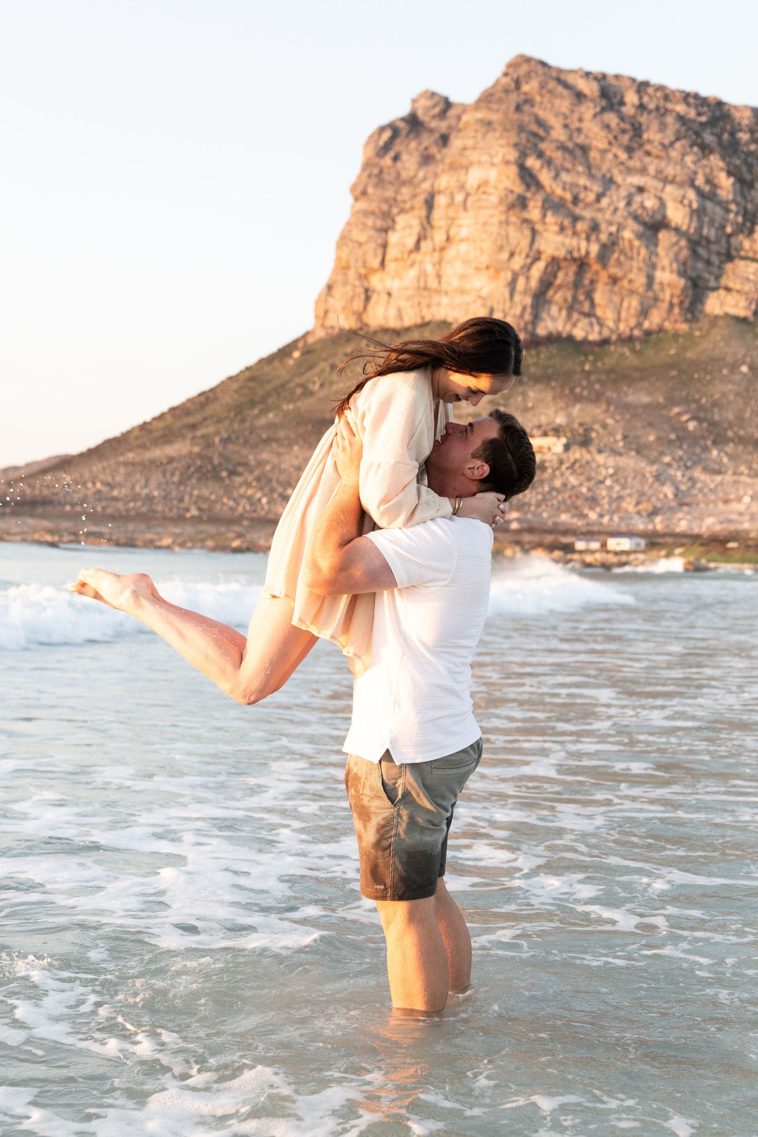 Playful couple embracing in shallow ocean water near scenic coastal cliff.