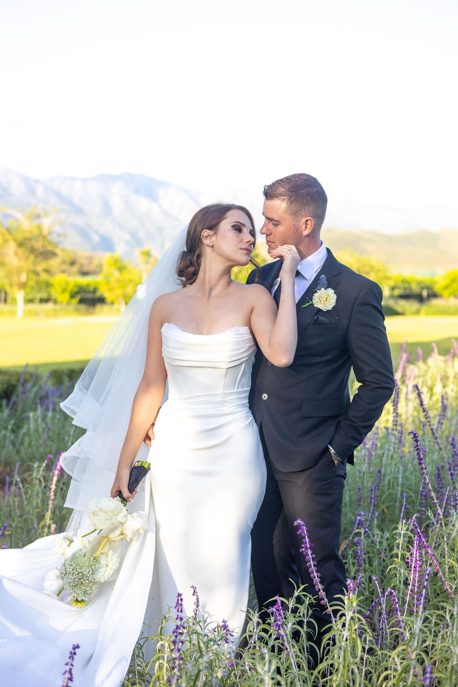 Elegant wedding couple embraces in field of purple wildflowers with mountains in background.