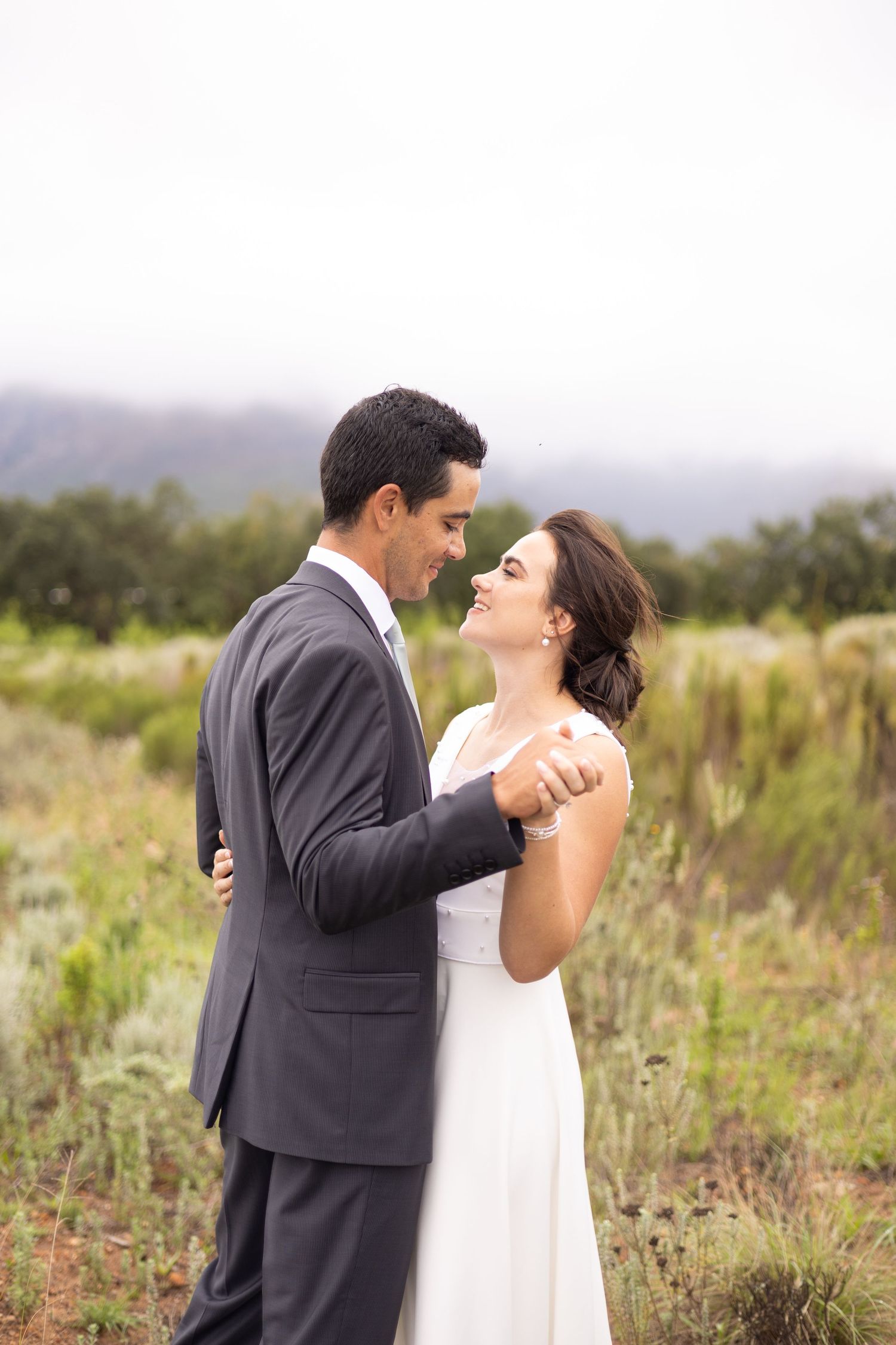 A couple in wedding attire embrace in a scenic outdoor meadow with mountains and overcast skies in the background.